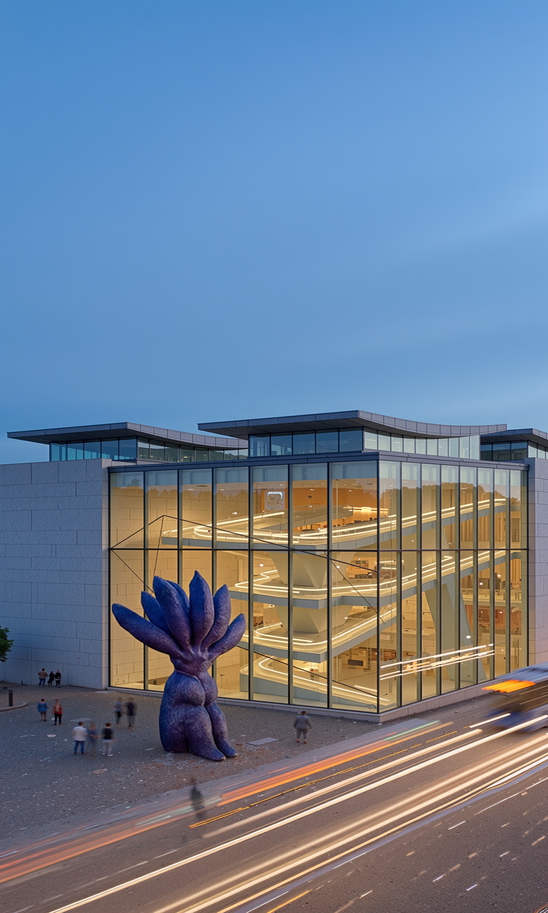 Twilight view of the museum with warmly illuminated spiraling ramps visible through the glass facade and the blue sculpture silhouetted against the evening sky