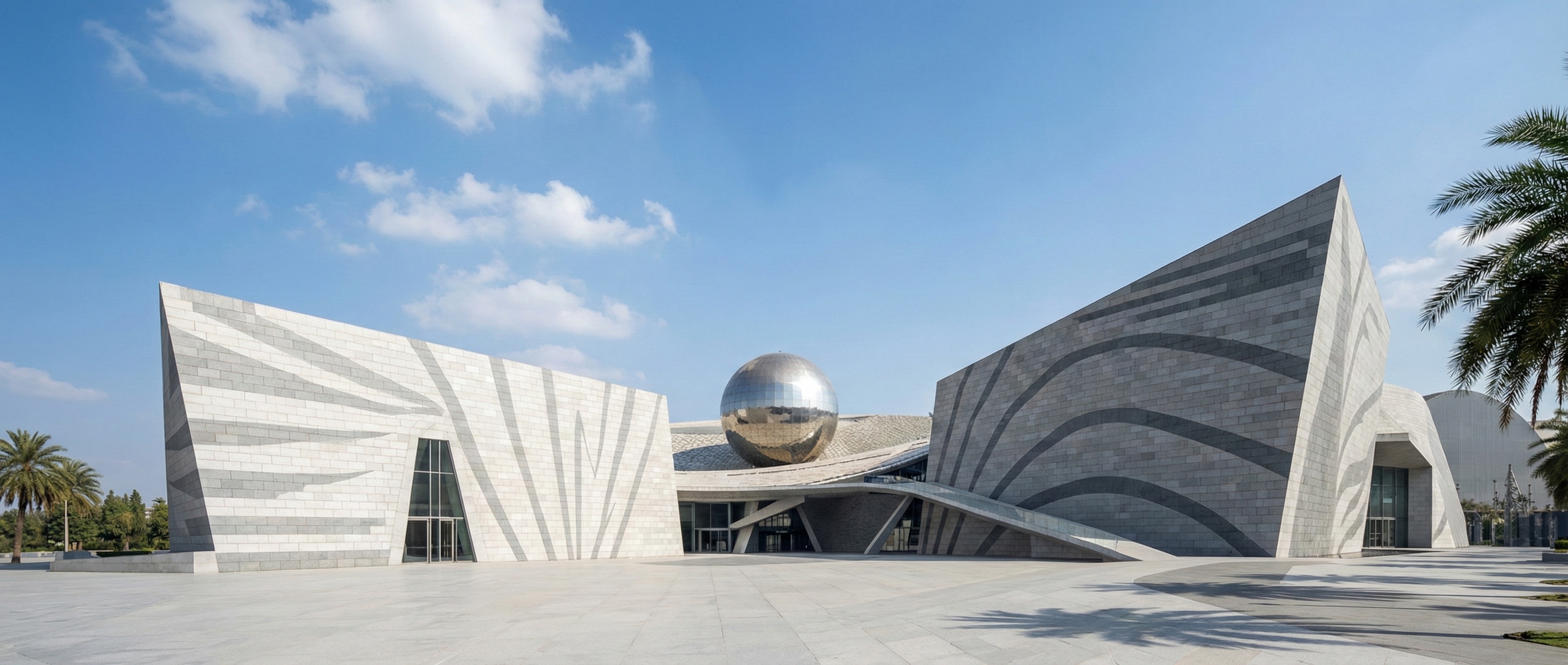 Twilight view of the Ambedkar Monument and Faith Center featuring two angular stone pavilions flanking a large reflective sphere, with visitors gathered in the expansive plaza