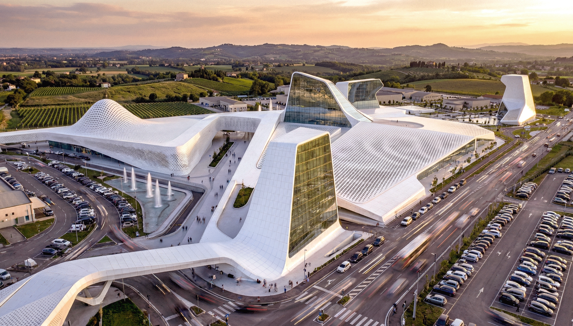 Aerial sunset view of the full Azzano complex with sculpted white rooflines, glass curtain wall towers, fountains, and the Italian countryside beyond