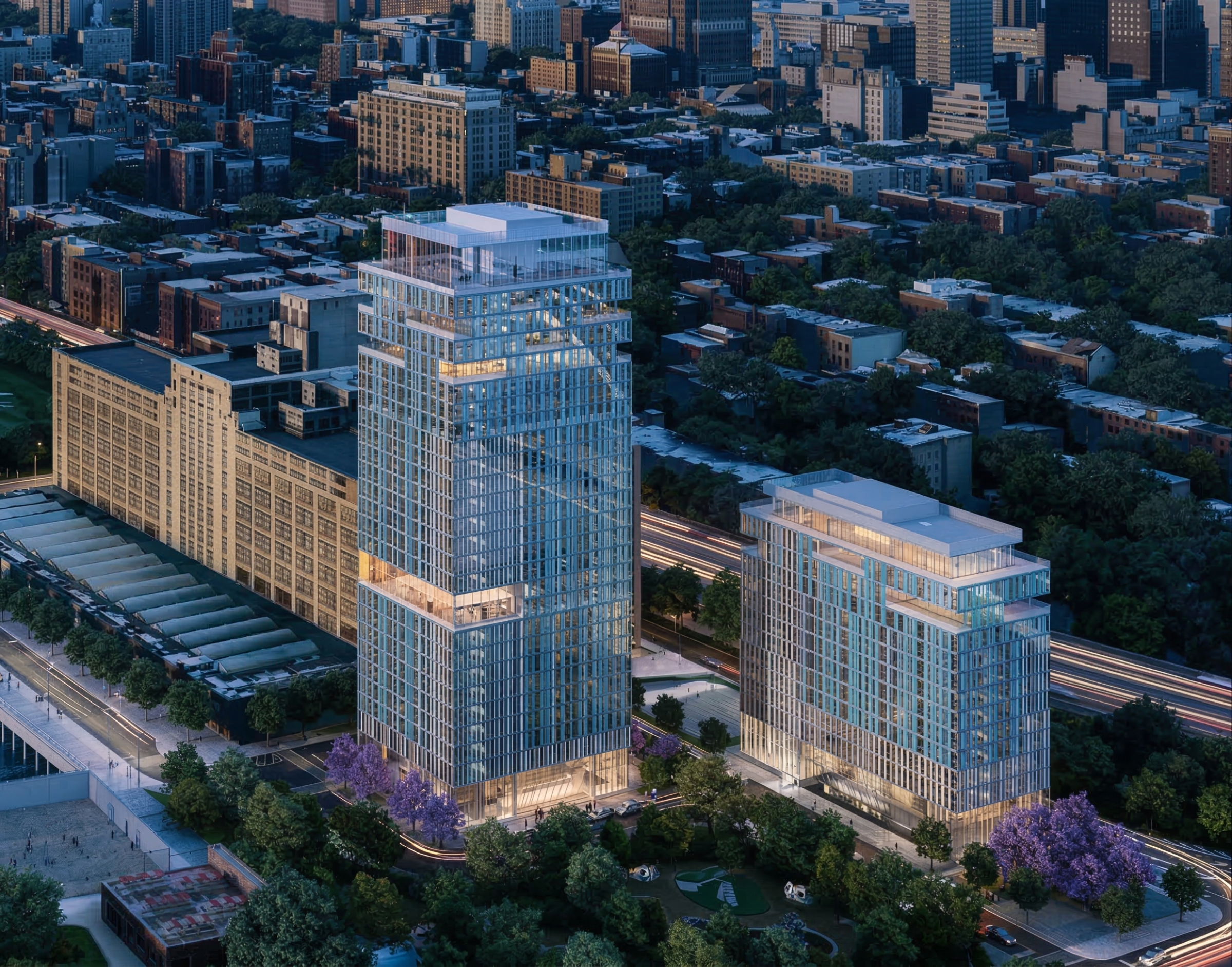 Aerial dusk view of the twin residential towers and mid-rise buildings with faceted glass facades, tree-lined streets, and the Brooklyn cityscape beyond