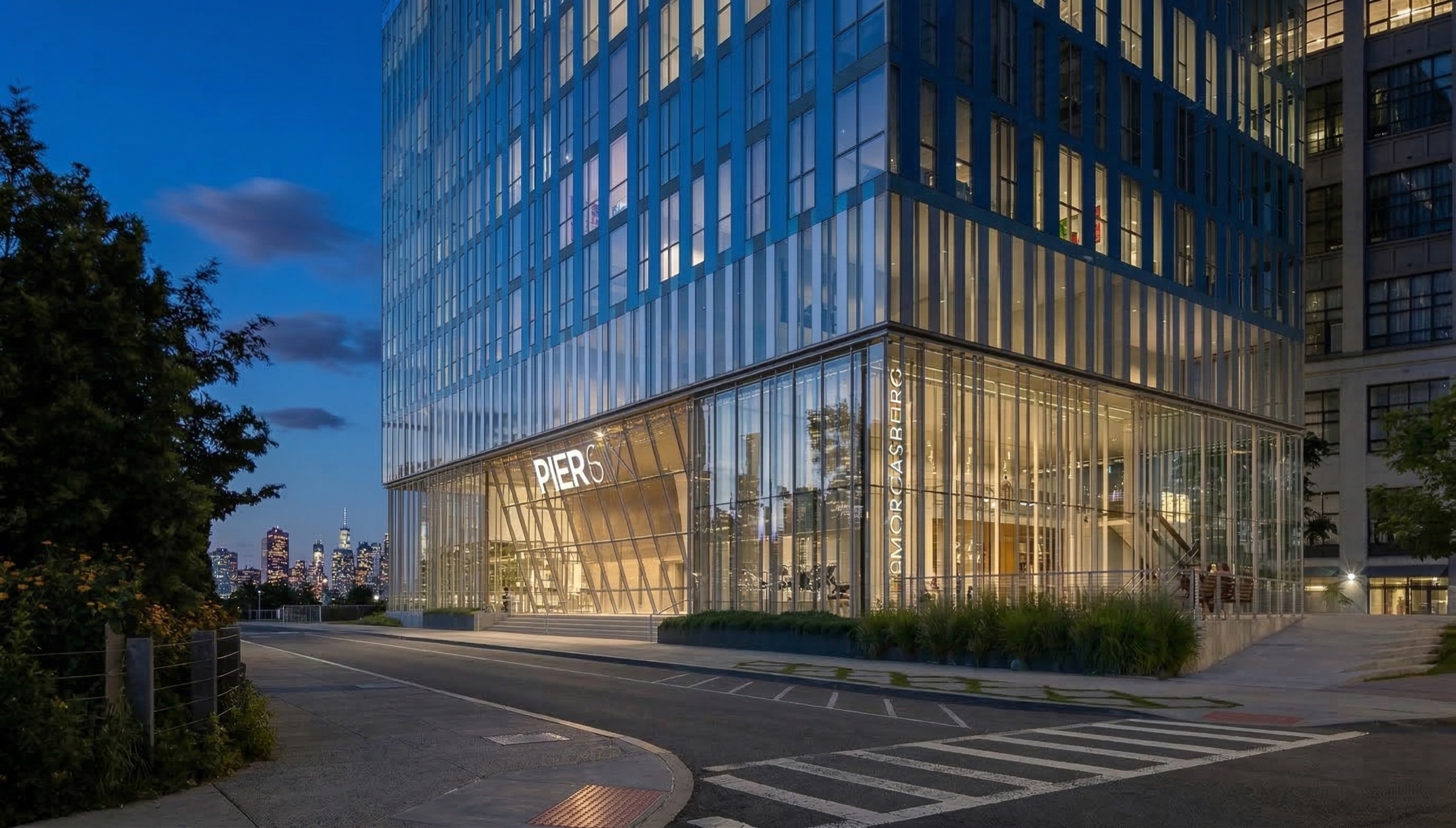 Evening corner view of the Pier 6 ground floor with illuminated double-height glass lobby, Pier 6 signage, landscaped perimeter, and Manhattan skyline visible beyond