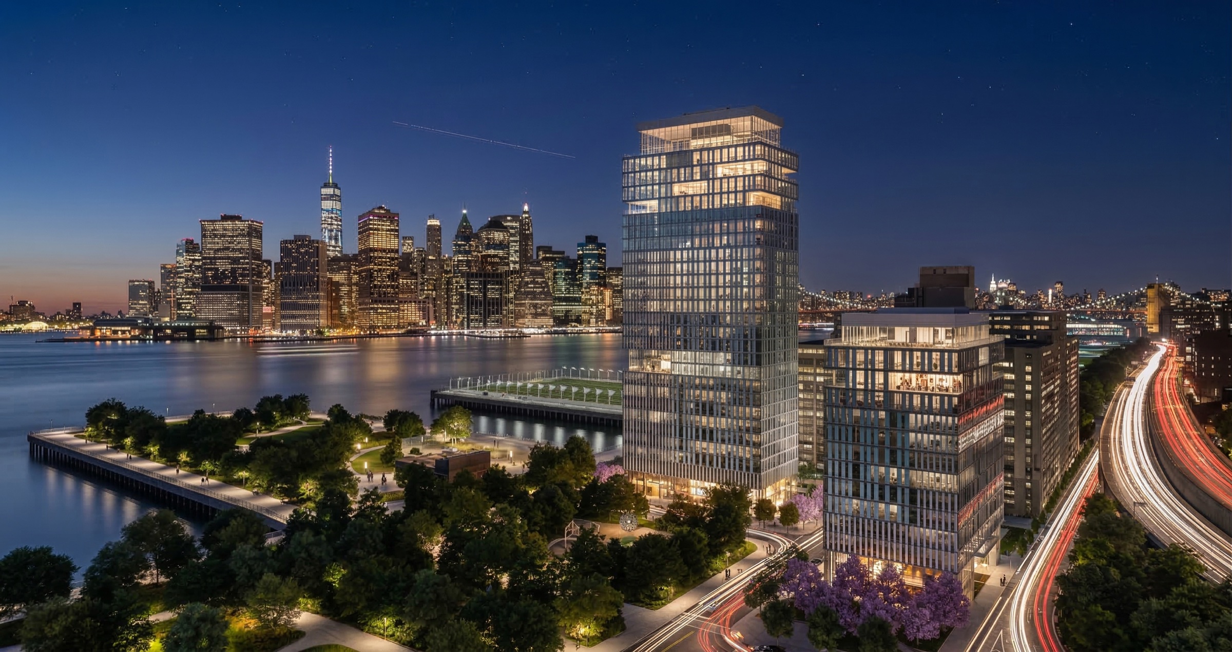 Panoramic night view of the Pier 6 tower illuminated against the Lower Manhattan skyline across the East River, with Brooklyn Bridge Park greenery and light trails on the roadway