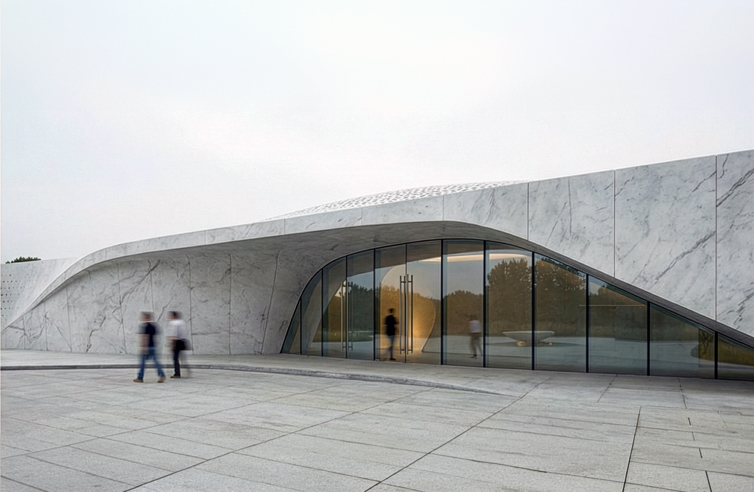 Beukenhof Crematorium entrance — curved marble canopy with full-height glazing and visitors approaching