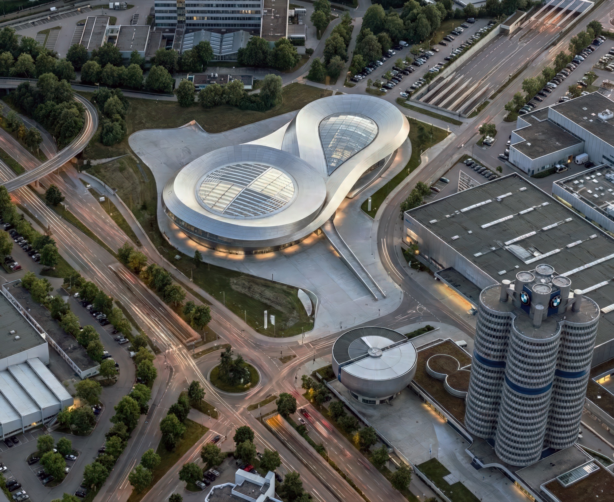 Aerial dusk view of the BMW Event and Delivery Centre showing the infinity-loop roof form alongside the BMW headquarters towers