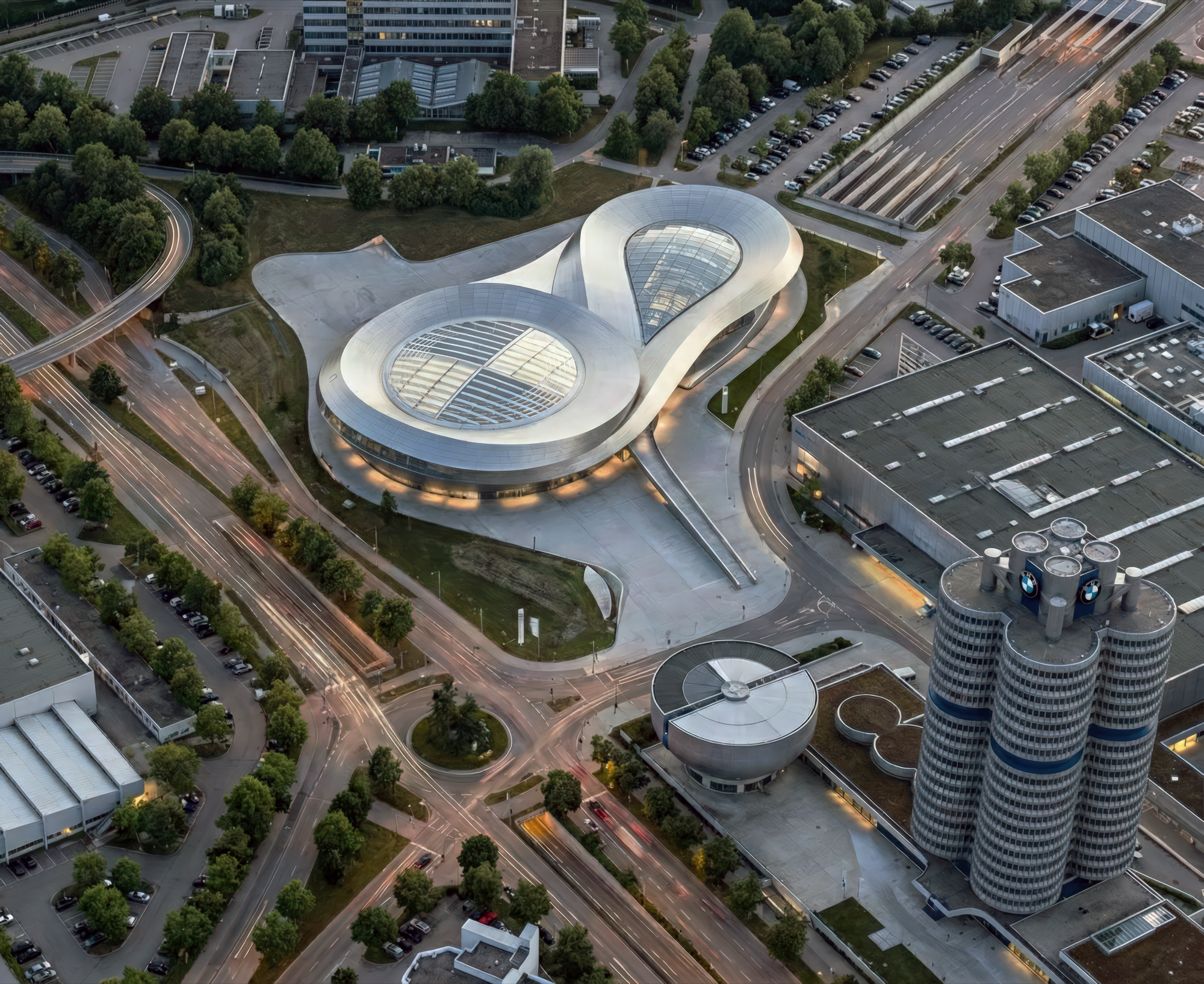 Aerial dusk view of the BMW Event and Delivery Centre showing the infinity-loop roof form alongside the BMW headquarters towers