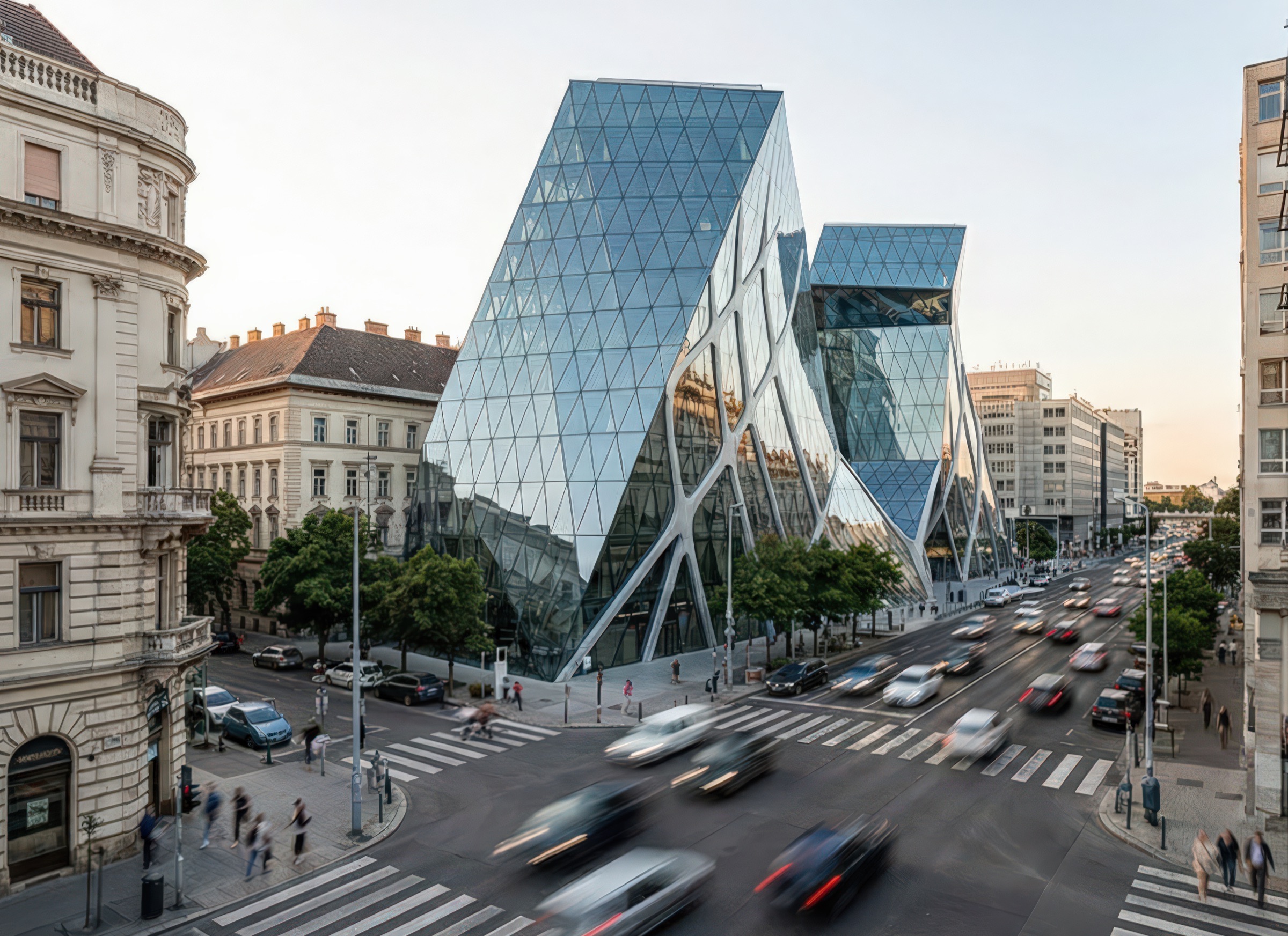 Elevated corner view of the Budapest Bank Tower at golden hour showing its angular glass volumes with diamond structural grid, adjacent historic buildings, a busy intersection with motion-blurred traffic, and mature street trees