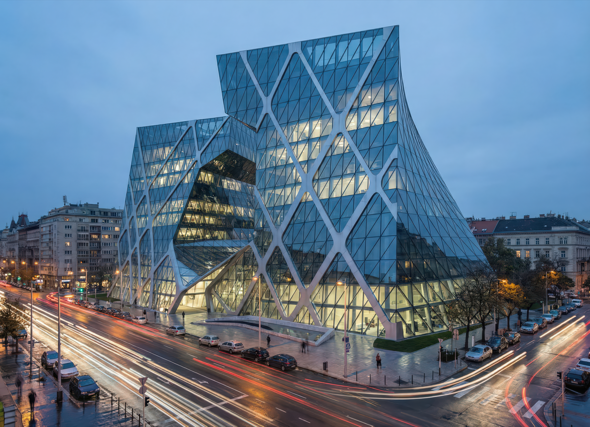 Elevated dusk view of the Budapest Bank Tower illuminated from within, its angular diamond-grid glass facade glowing against the historic Budapest streetscape with traffic light trails on the boulevard below