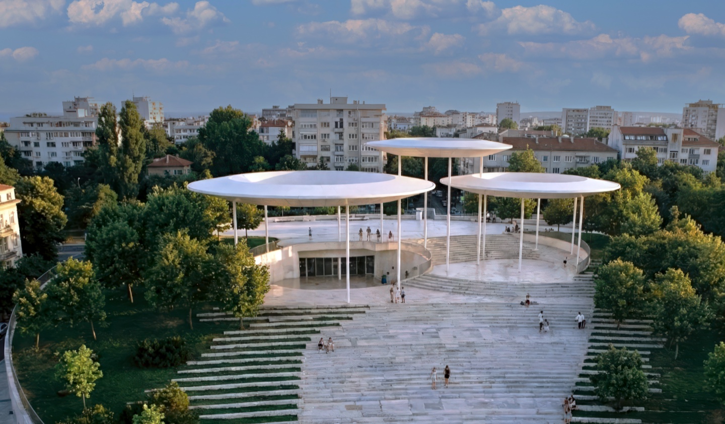 Elevated view of the center showing the disc-shaped shade canopies atop terraced stone steps with the city beyond