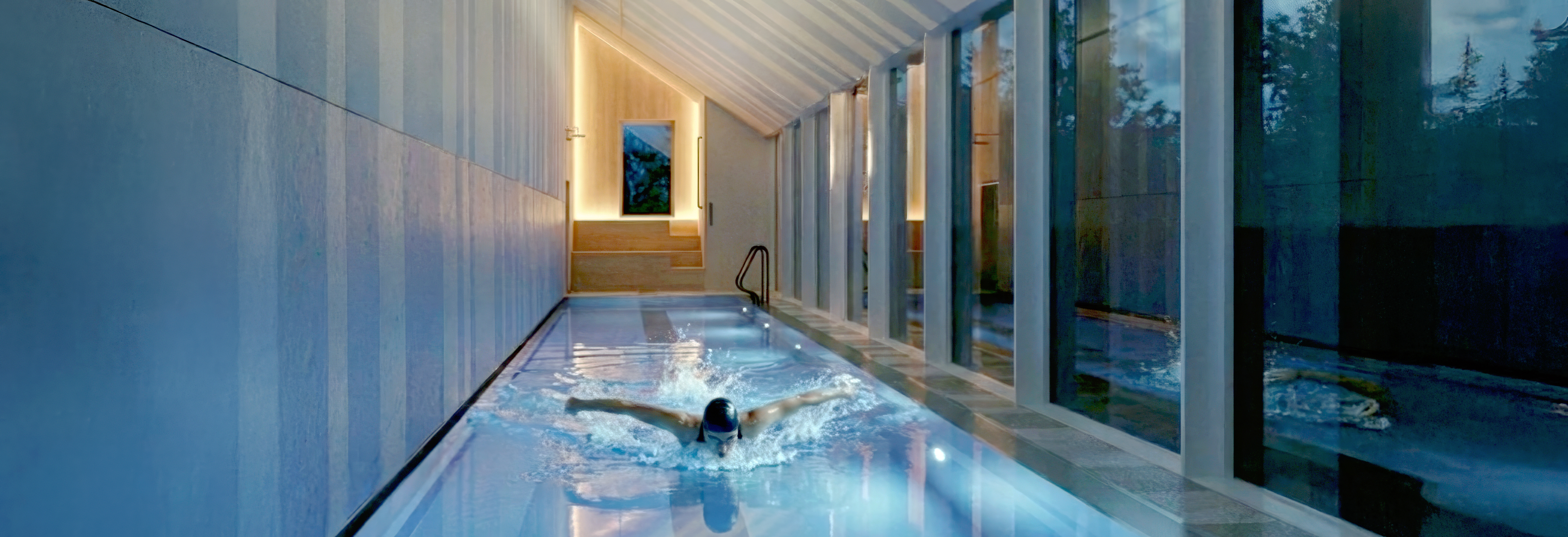 Interior dusk view of the Butterfly Pool Pavilion with a swimmer performing the butterfly stroke in the lap pool, the pitched striped ceiling warmly uplit above and full-height glazing on the right opening to the wooded landscape