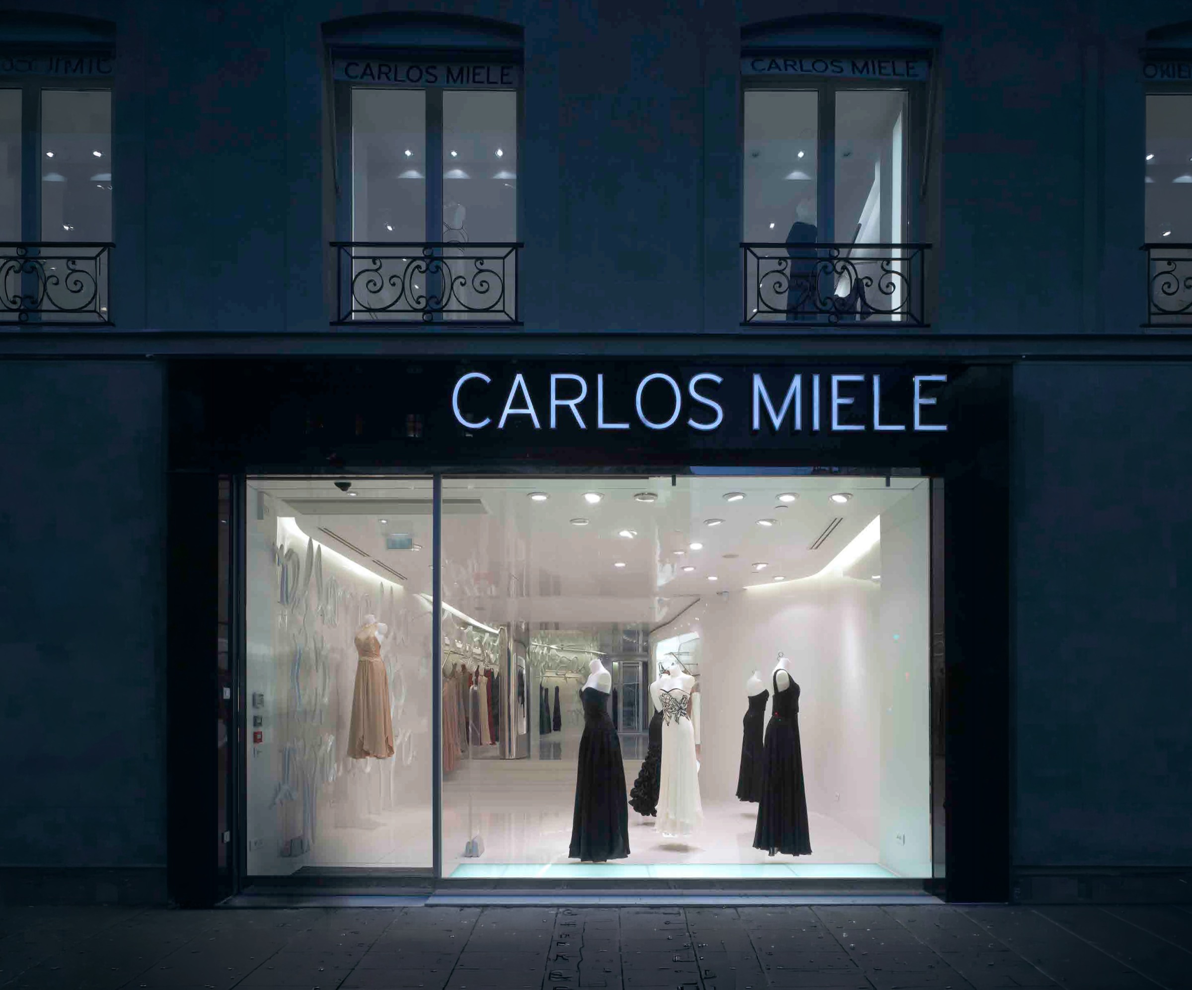 Carlos Miele Flagship Store exterior at night on Rue Saint-Honoré, Paris, with illuminated storefront window displaying mannequins in evening gowns beneath the CARLOS MIELE signage and Parisian balconies above