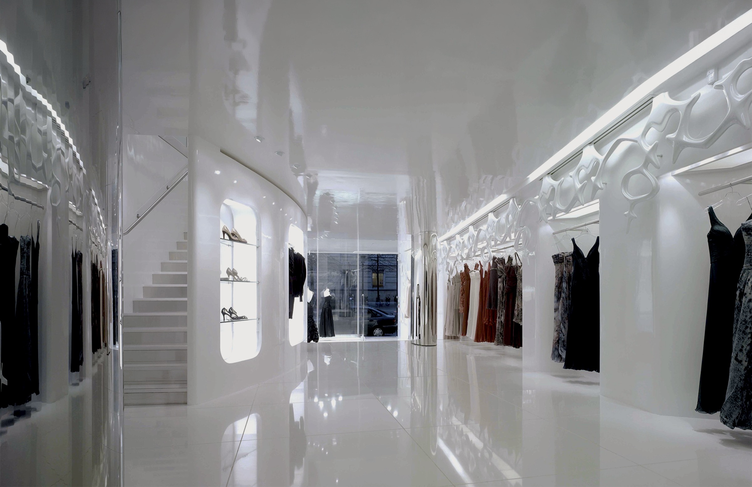 Main corridor with high-gloss white surfaces, organic fiberglass wall sculpture, integrated staircase with illuminated shoe display, and garments displayed along both sides