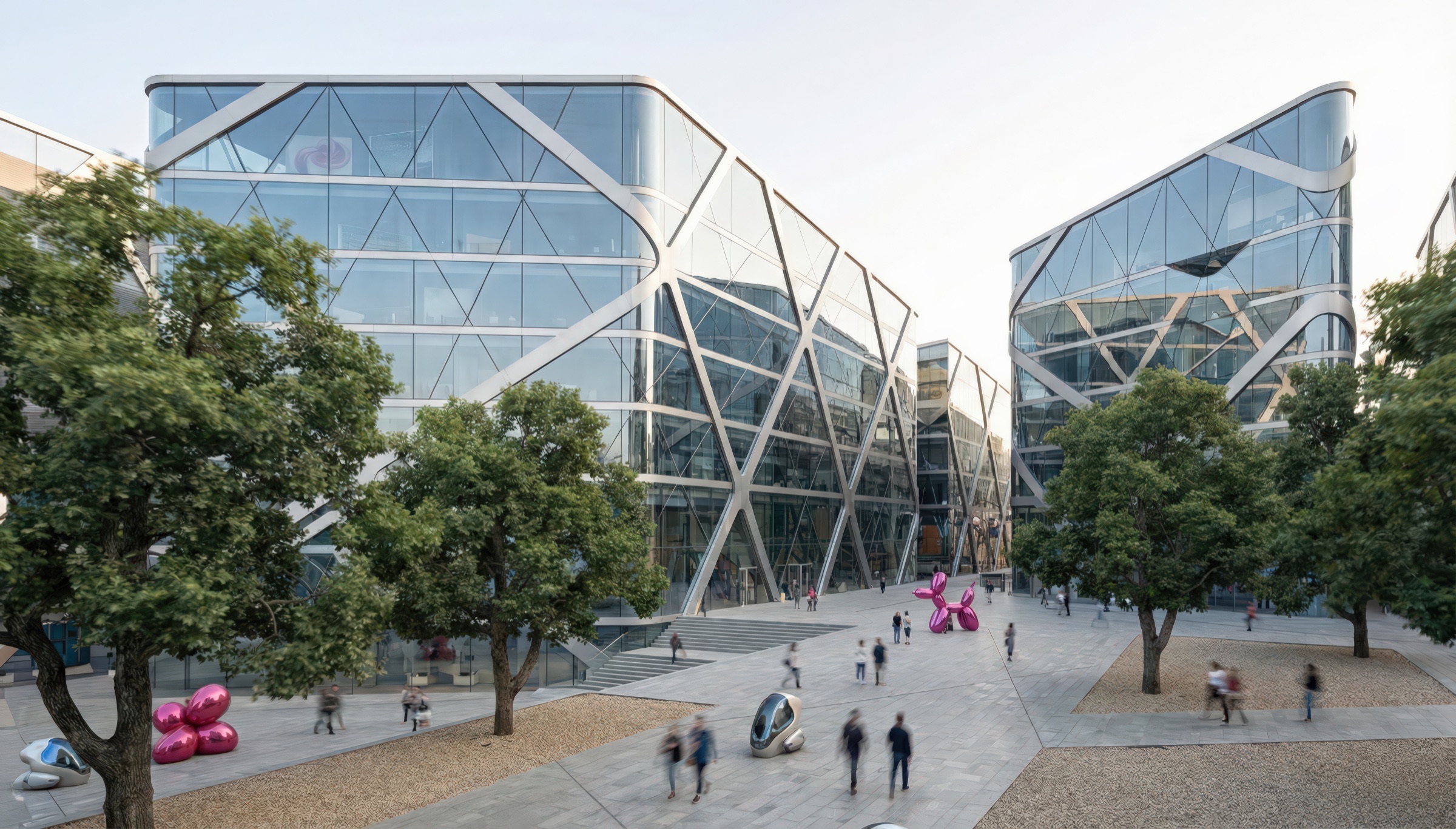 Street-level view of the tech campus plaza with pedestrians walking beneath mature trees, two faceted diagrid glass buildings with angular facades on either side, and public art installations