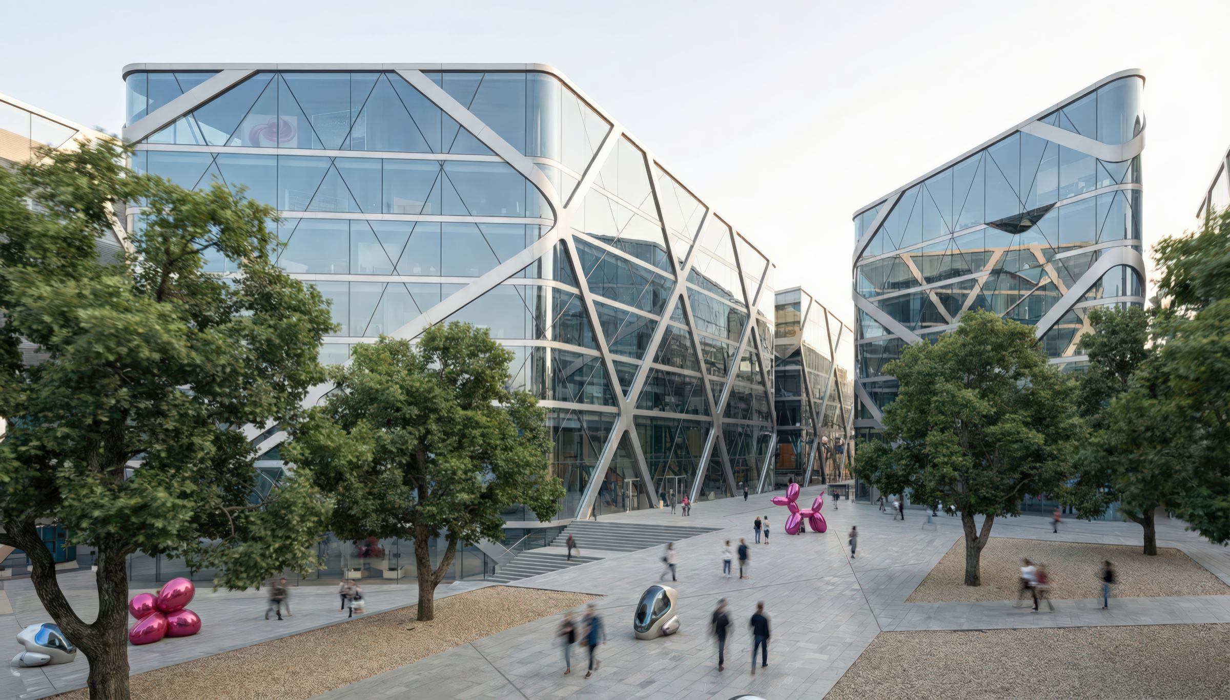 Street-level view of the tech campus plaza with pedestrians walking beneath mature trees, two faceted diagrid glass buildings with angular facades on either side, and public art installations