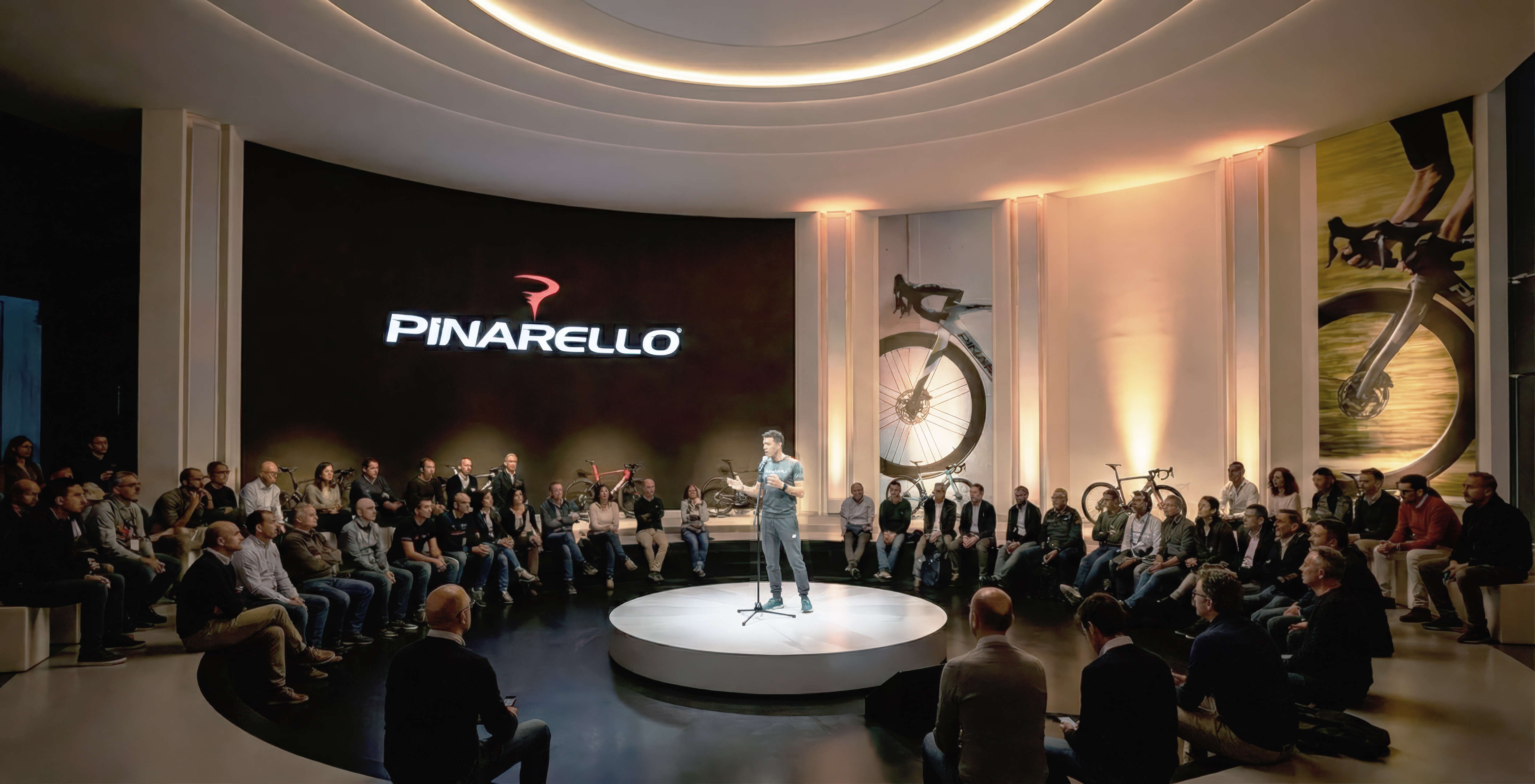Brand presentation space with a presenter on a circular central platform addressing a seated audience under a curved LED wall bearing the Pinarello logo