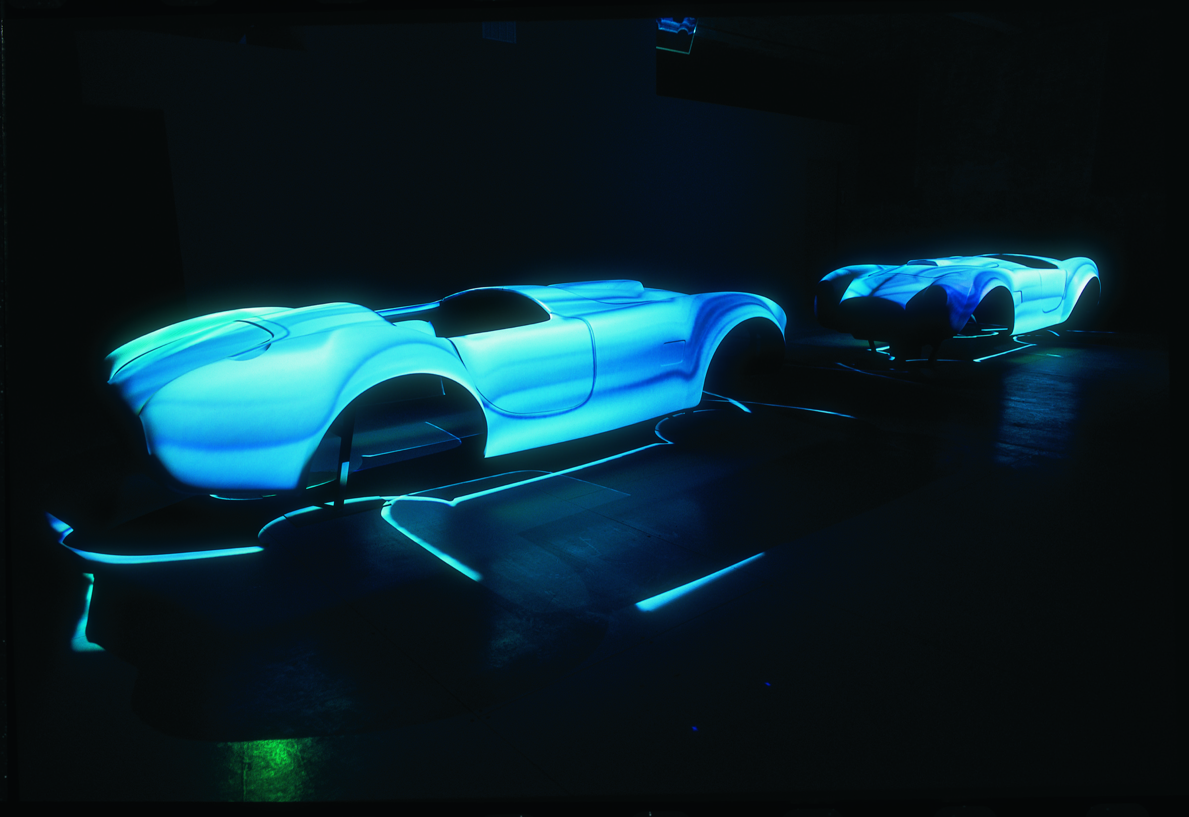 Two Cobra race car body forms illuminated with cyan-blue projected light in a darkened space, glowing neon lines tracing paths across the floor