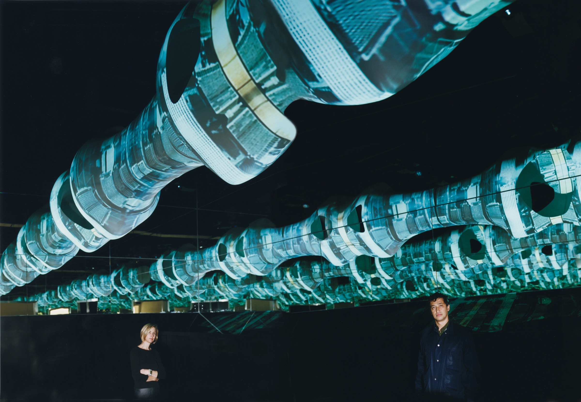 Hani Rashid and Lise Anne Couture beneath the Fluxspace 3.0 installation at Documenta XI — biomorphic spines suspended from the ceiling with projected cityscapes
