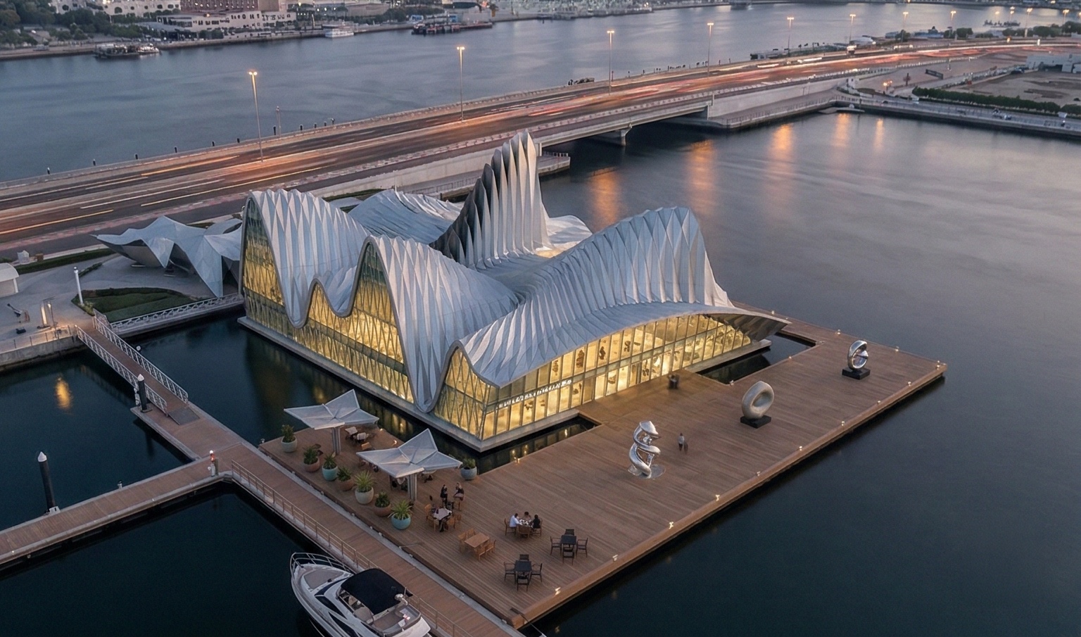Aerial twilight view of the Dubai Contemporary Arts Center illuminated from within, situated on a waterfront pier with surrounding deck terraces and the city bridge beyond