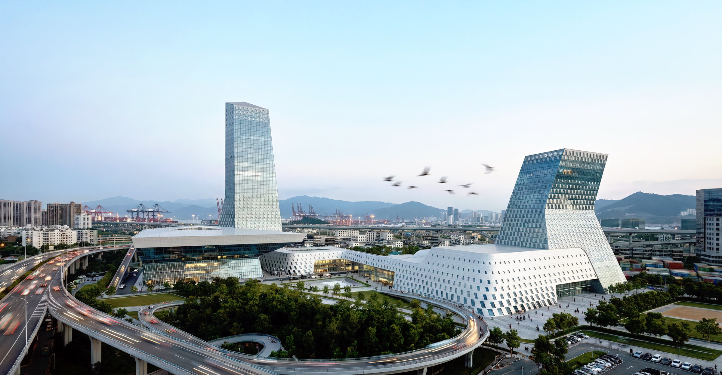 Aerial view of the Emerald Island complex showing the glass tower, angular performing arts center, and waterfront campus with birds in flight over the harbor