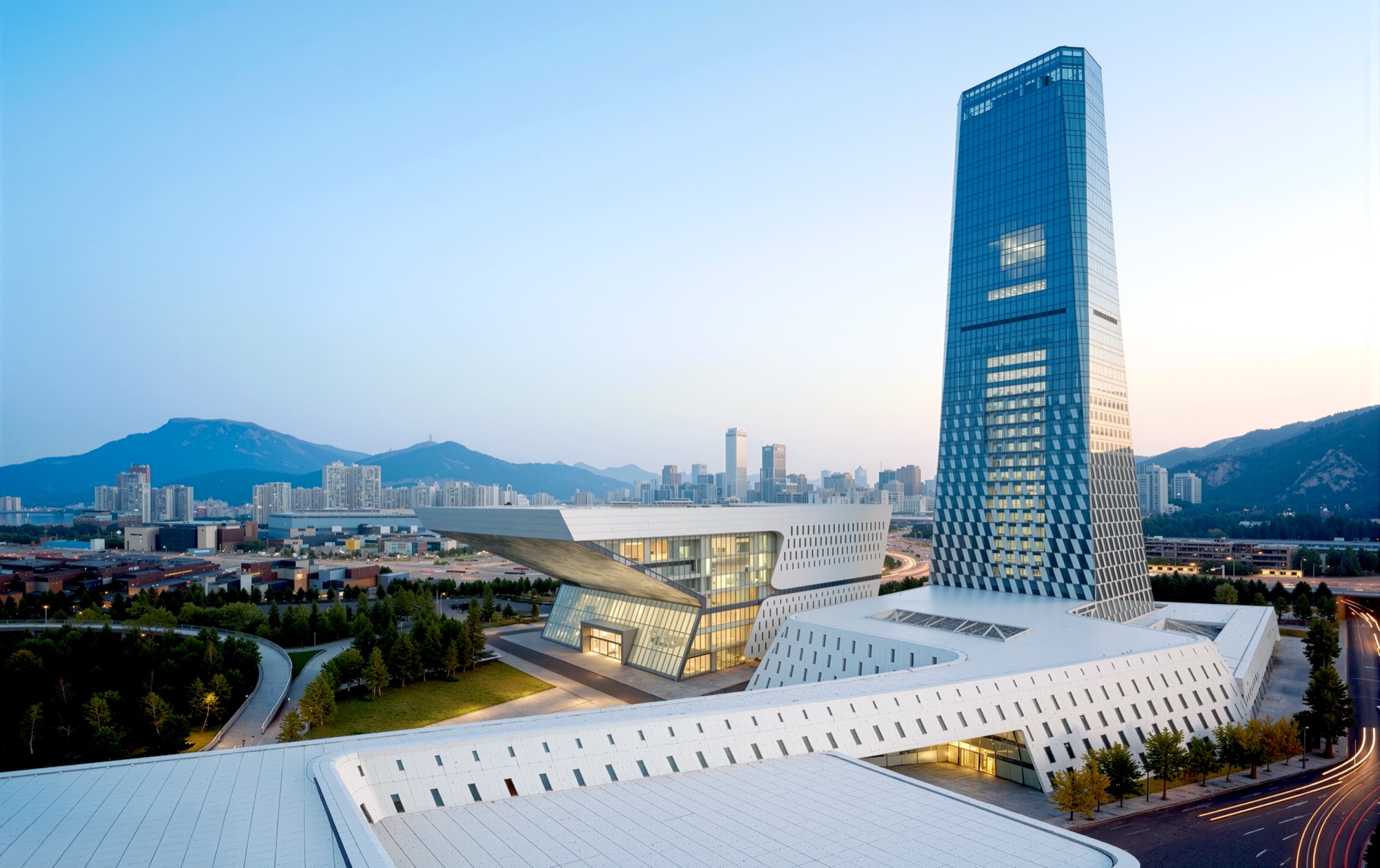 Dusk aerial showing the performing arts center's cantilevered volumes and the illuminated tower with city skyline and mountains beyond