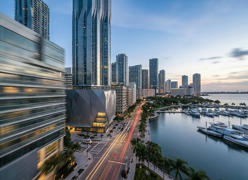 Elevated view of the Flagler Towers and their faceted podium along Biscayne Boulevard with the marina and bay visible in the background at dusk