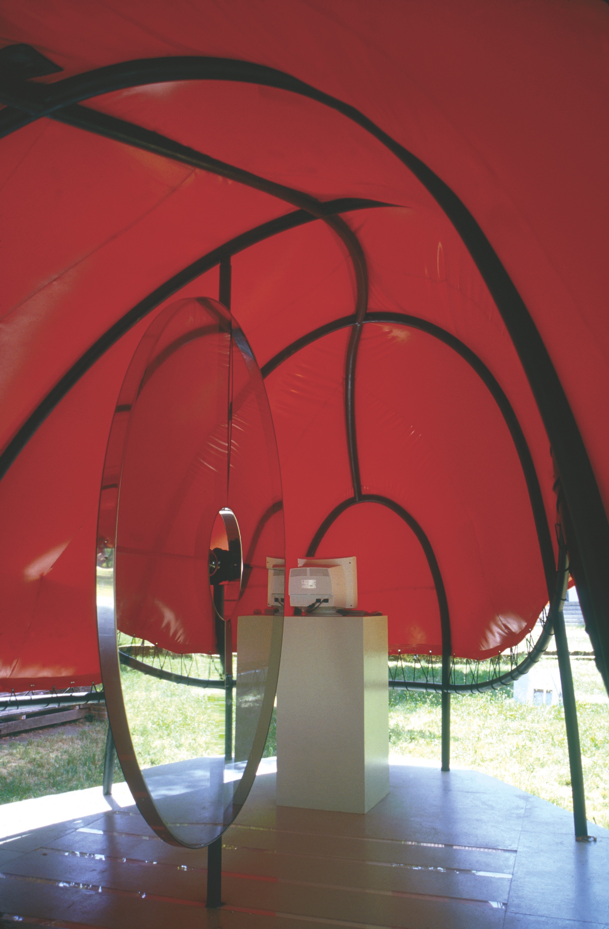 Interior of the Flux Pavilion with circular mirrored elements and a projection pedestal — red canopy above, wooden deck below