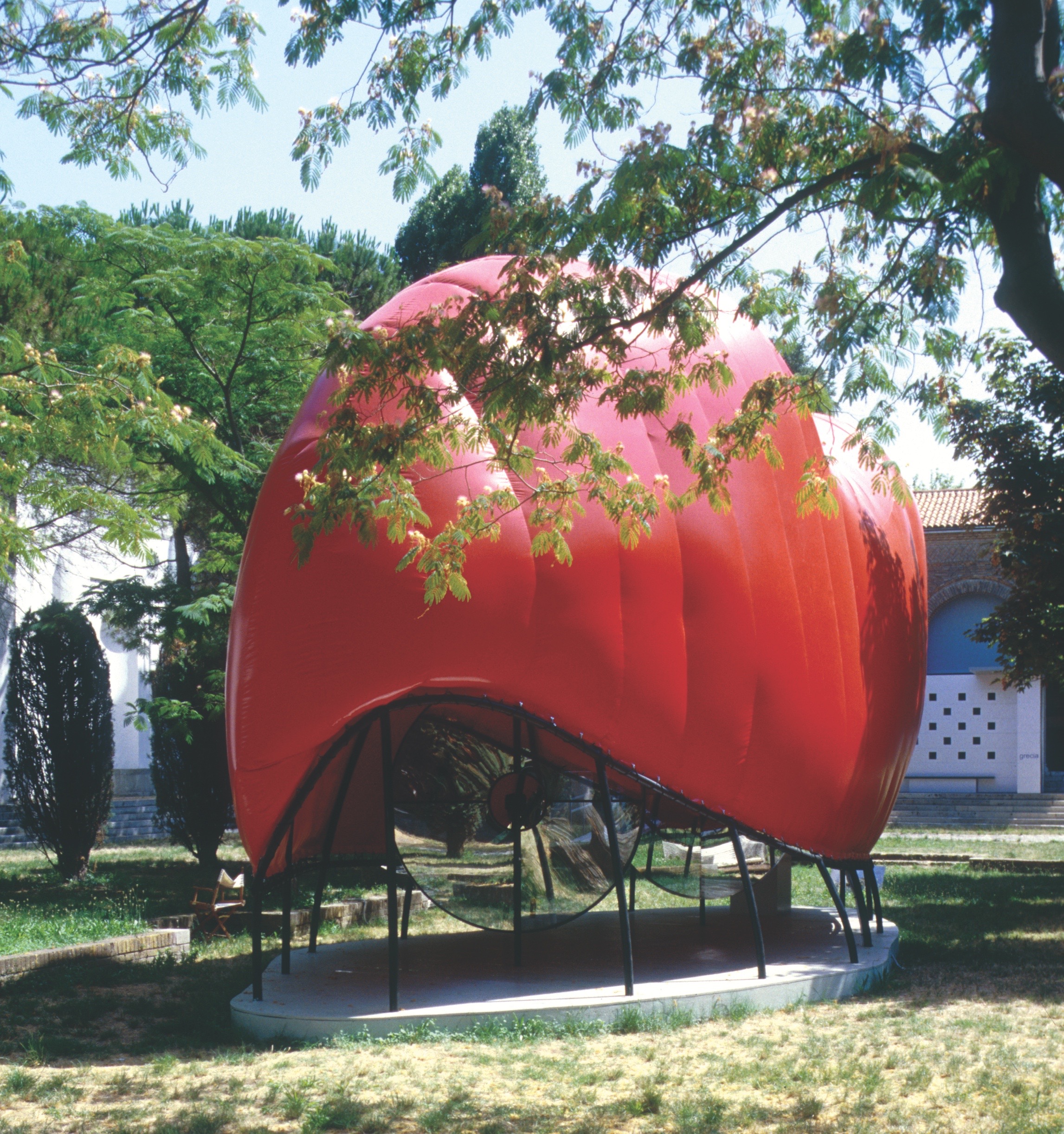 Side view of the Flux Pavilion — the biomorphic red inflatable envelope supported on curved steel armatures among the Giardini trees