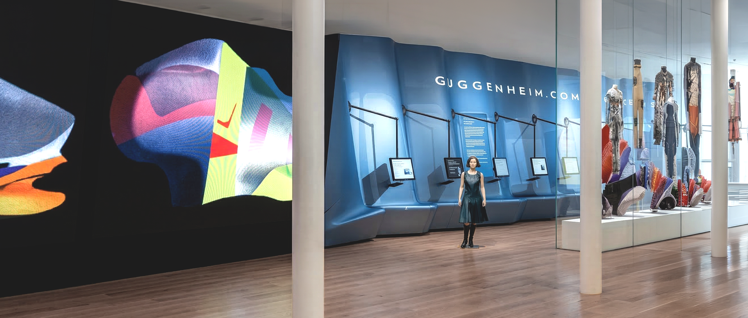 Interior entry zone with a curved blue-lit wall branded GUGGENHEIM.COM, articulated arm-mounted display screens, fashion vitrines, and a large-scale digital art projection