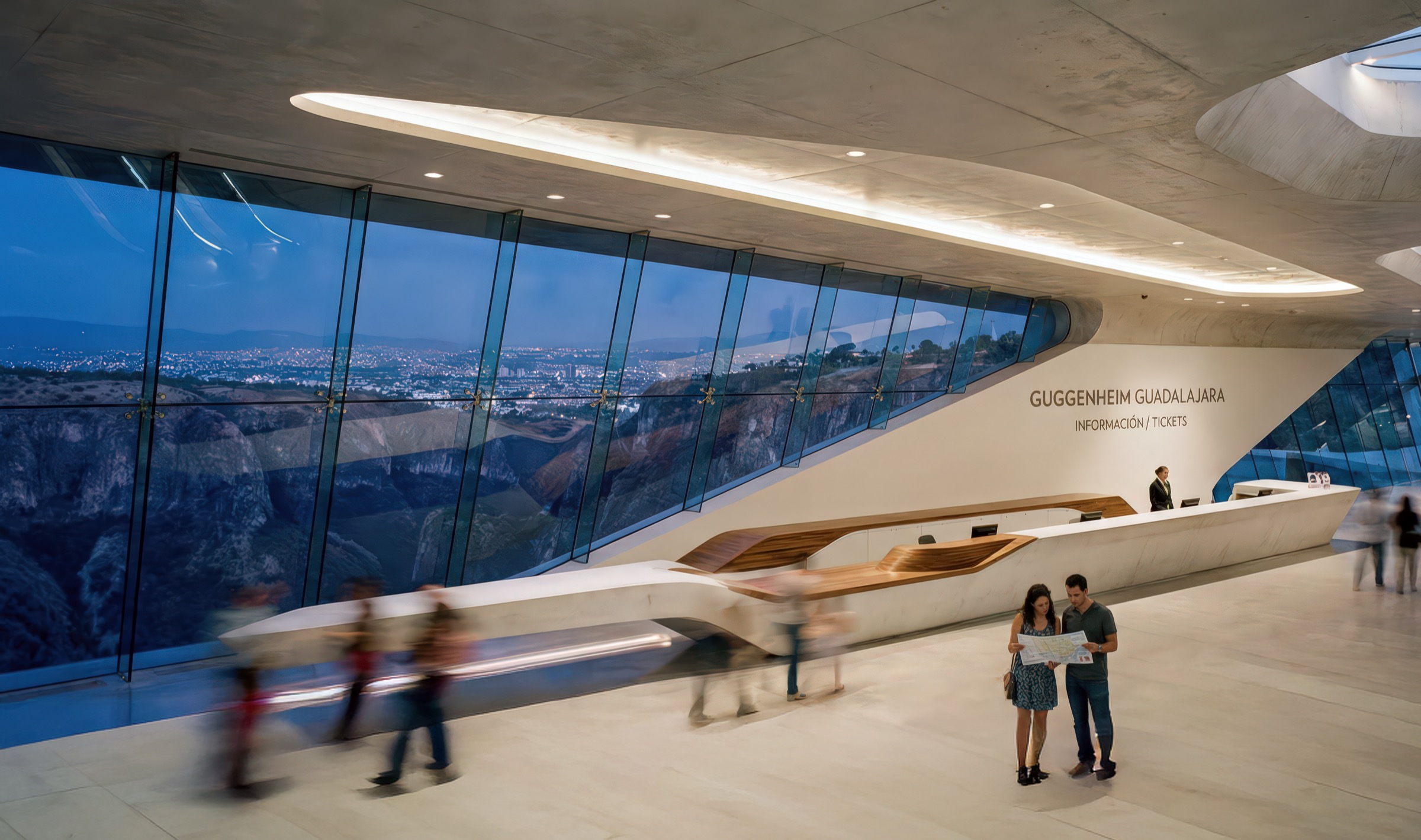 Ticketing lobby of the Guggenheim Guadalajara with GUGGENHEIM GUADALAJARA signage on a sweeping white reception desk with wood accent, angled floor-to-ceiling glass walls framing views of the Barranca canyon and city skyline at dusk, and motion-blurred visitors
