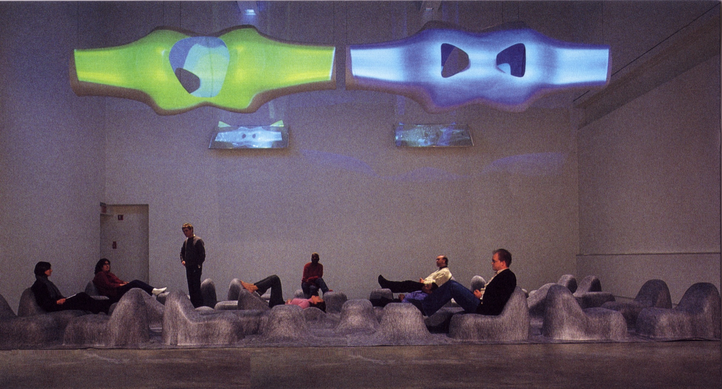 Wide view of the Stratascape installation at ICA — visitors reclining on the grey felt landscape beneath two glowing biomorphic M-Scape forms, one green and one blue