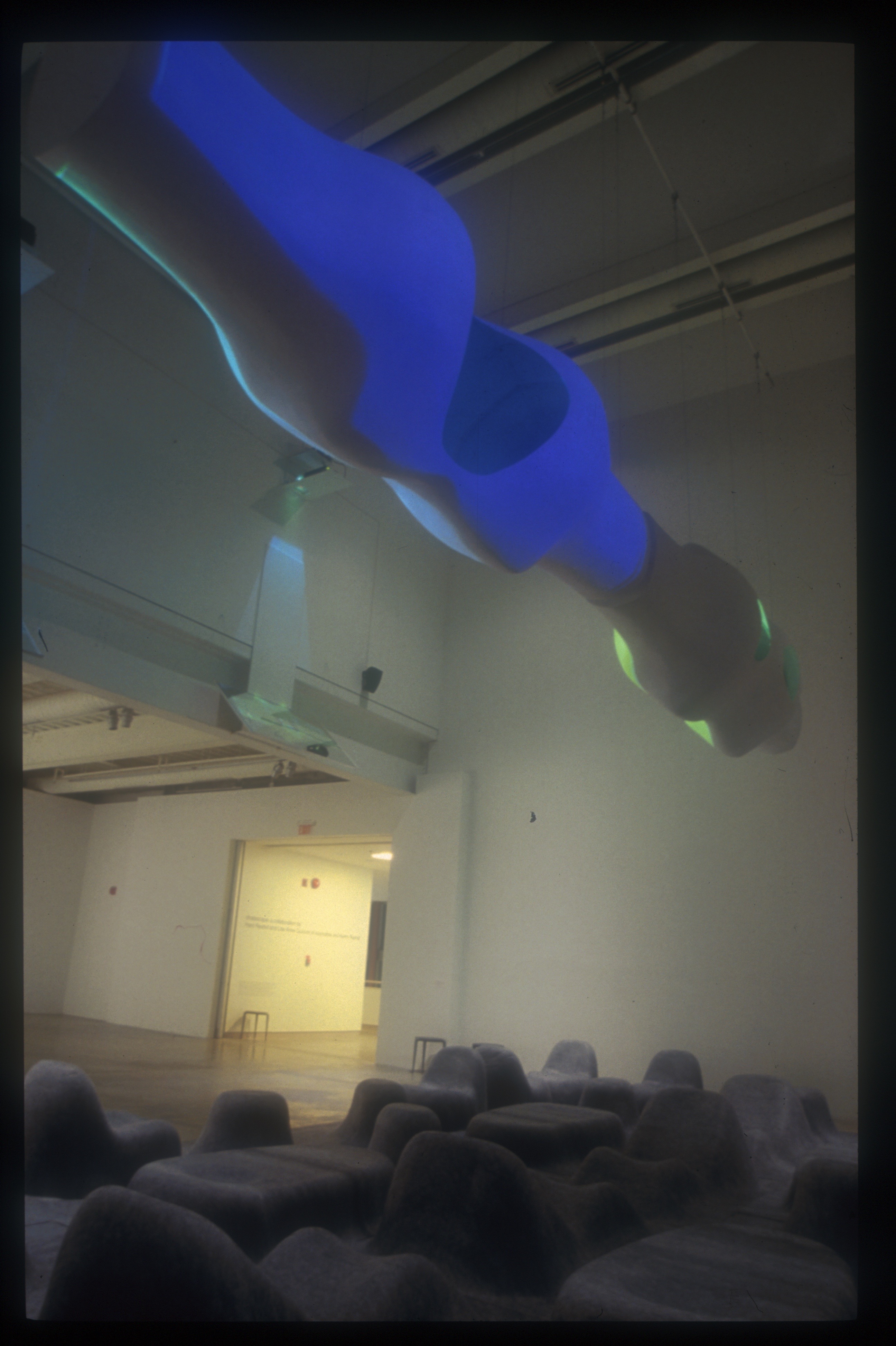 Vertical view looking up at a suspended biomorphic form glowing deep blue — the M-Scape hovering above the gallery space
