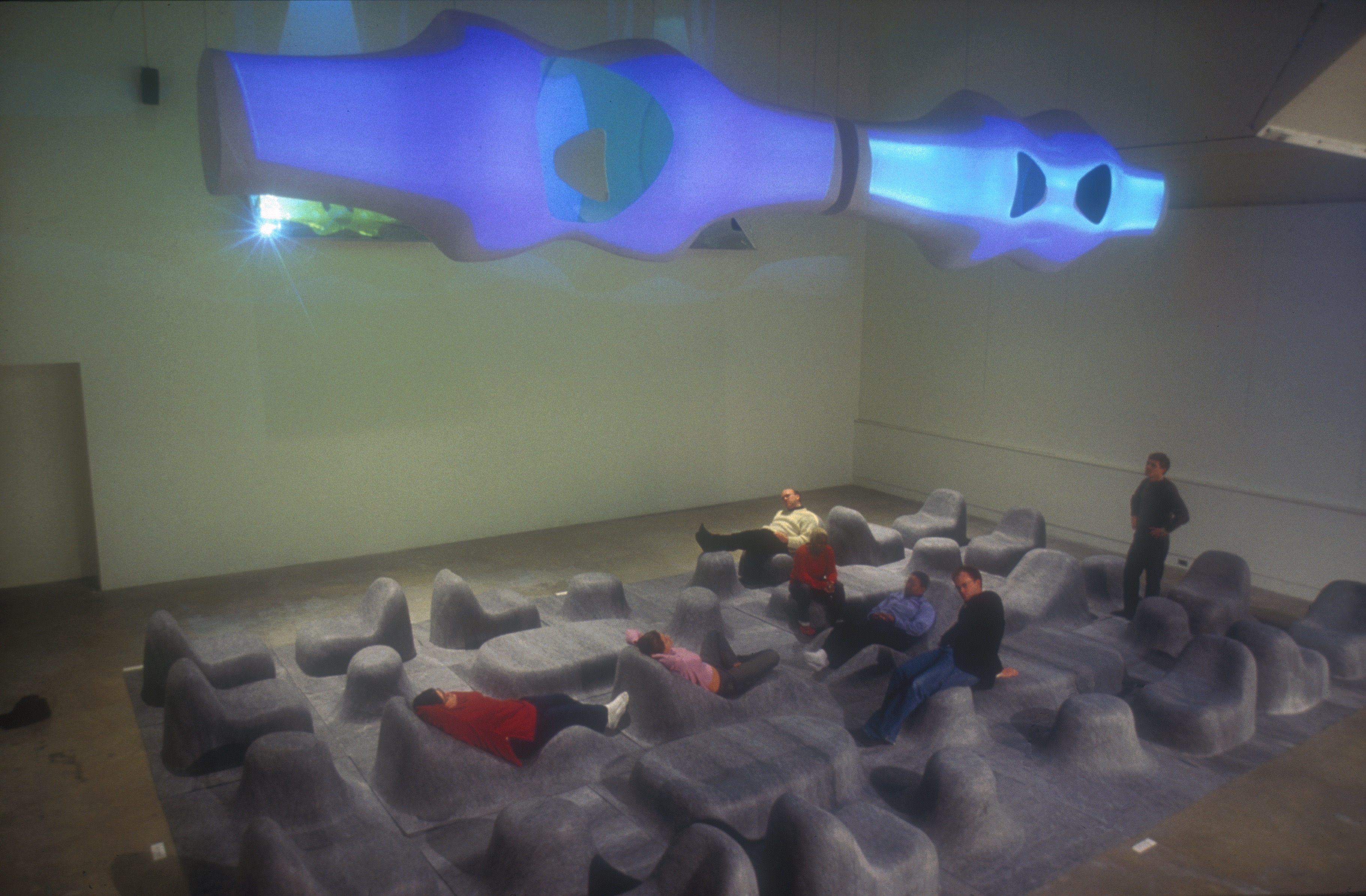Overhead view of the installation — visitors scattered across the undulating grey felt landscape with illuminated blue forms above