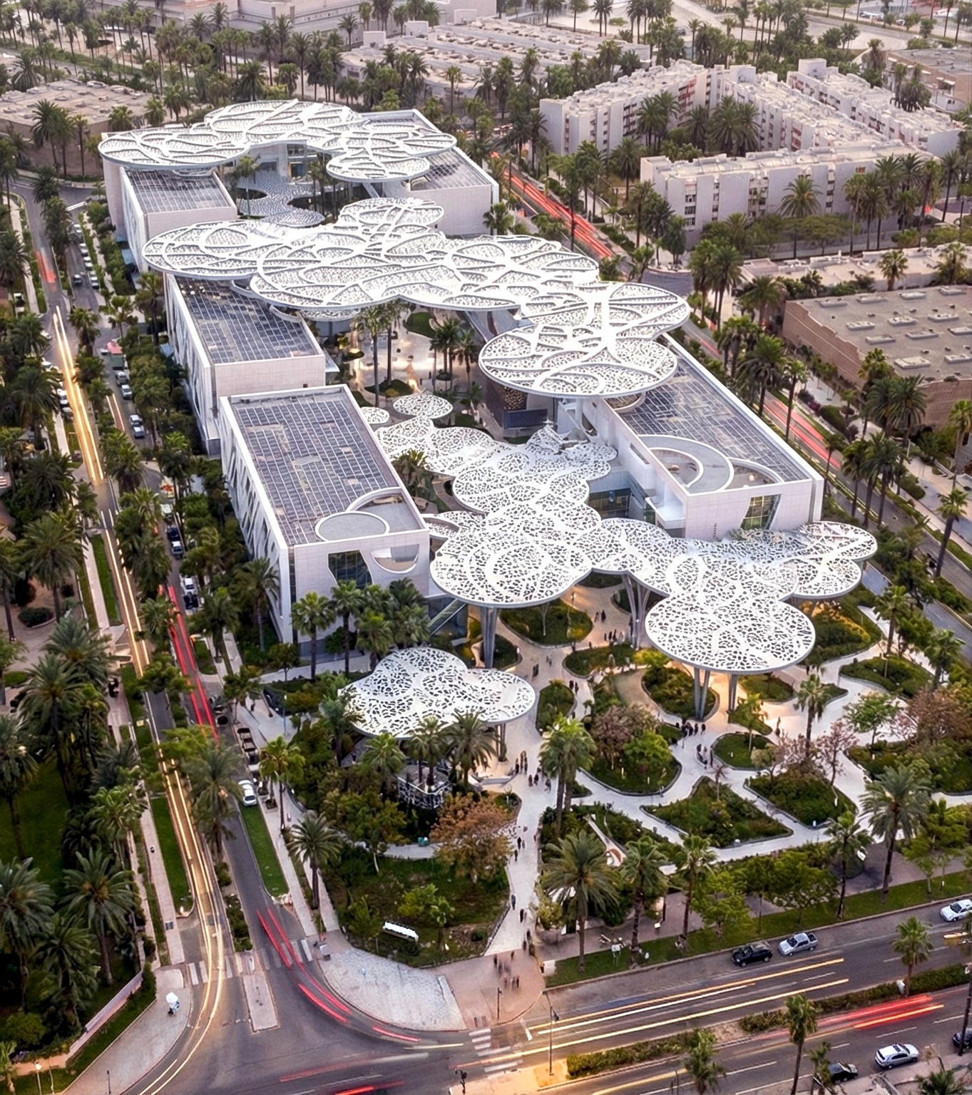 Angled aerial view of the campus — perforated lace canopies hovering over courtyards and gardens, surrounding streets and residential blocks visible at the edges