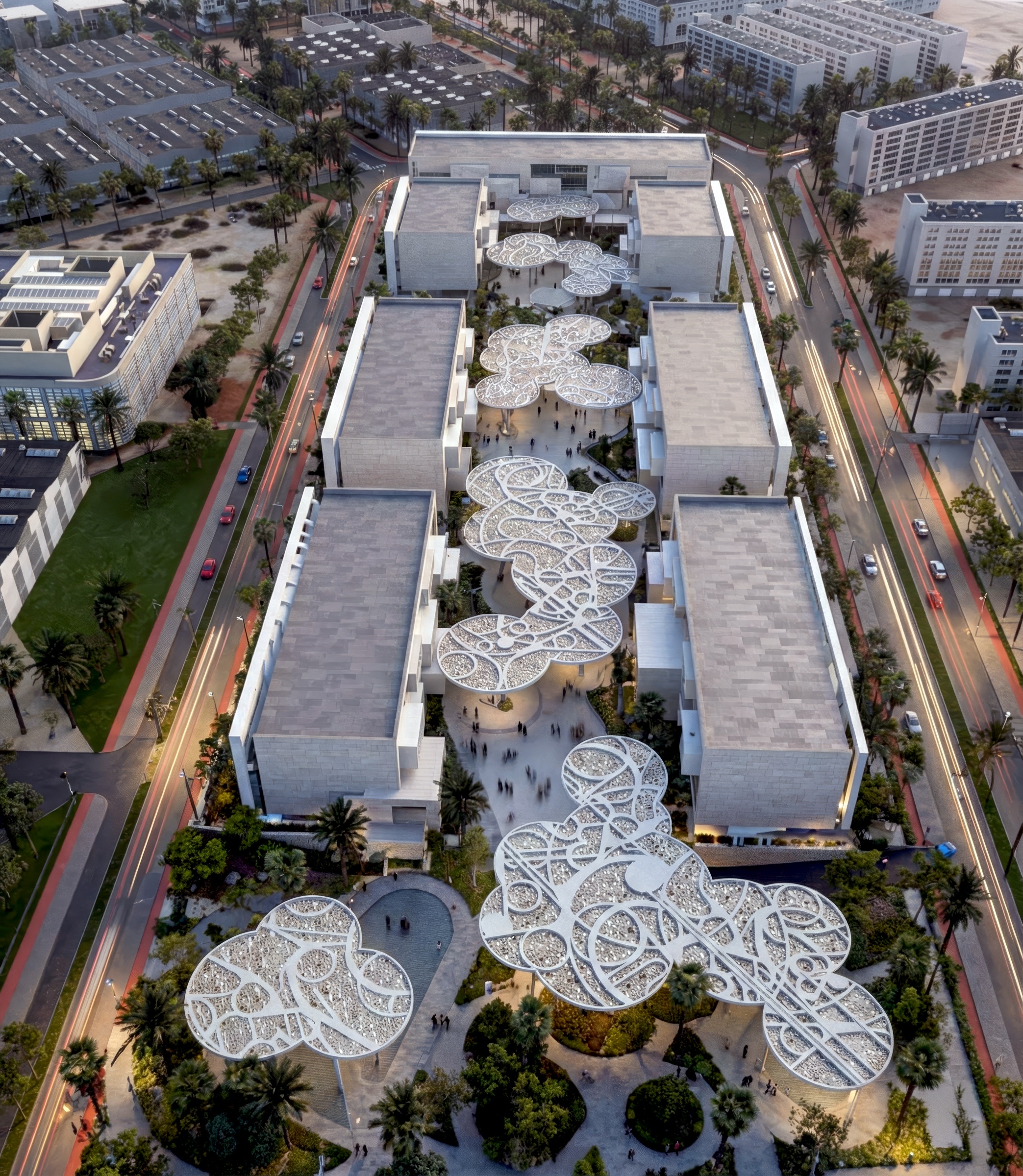 Top-down aerial view of the campus — perforated lace canopies arrayed across a rectilinear plan, gardens threaded between pavilions, surrounding streets and residential blocks visible at the edges