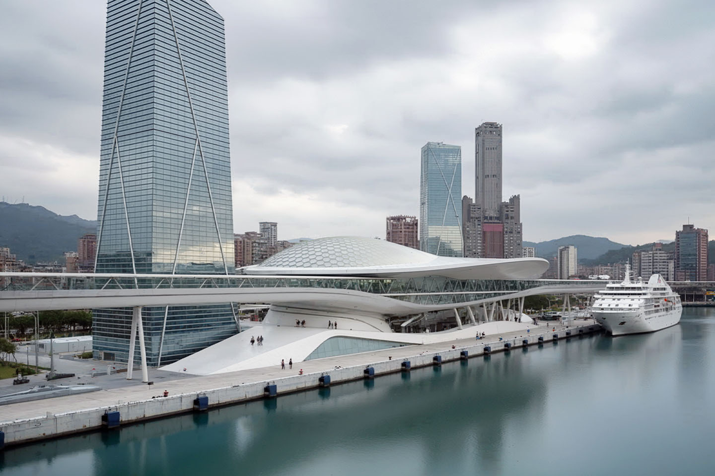 Aerial view of the full terminal complex from the harbor showing the hexagonal dome canopy, flanking tower, docked cruise ship, and Kaohsiung's mountainous skyline