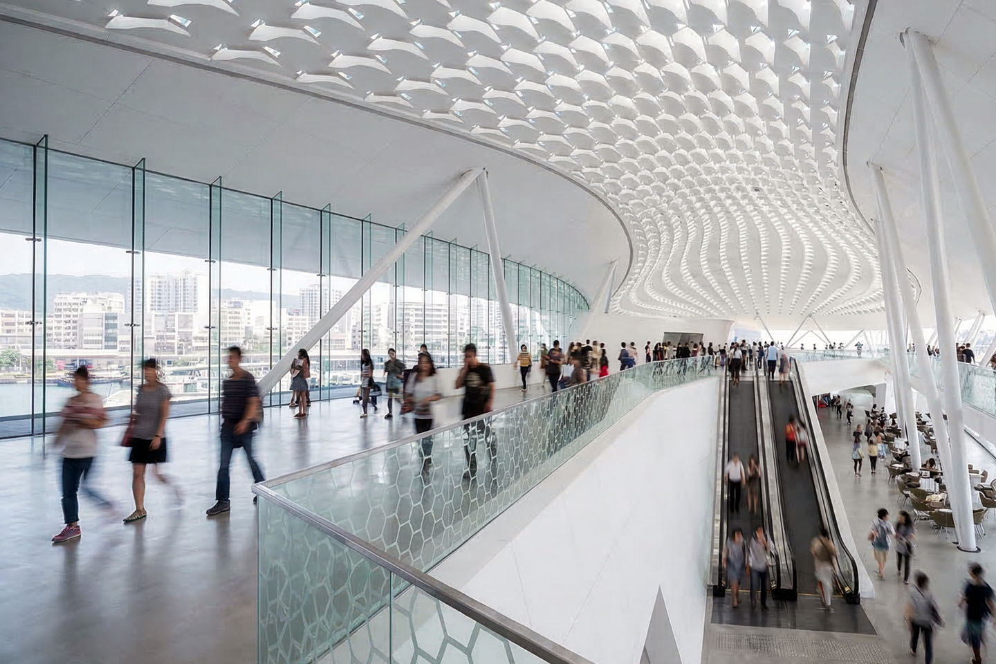 Upper-level concourse with undulating geometric ceiling pattern, curved glass wall overlooking the harbor, escalators, and pedestrians