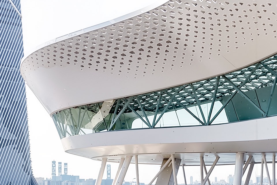 Detail of the perforated canopy roof with structural truss system visible beneath and tower rising beyond