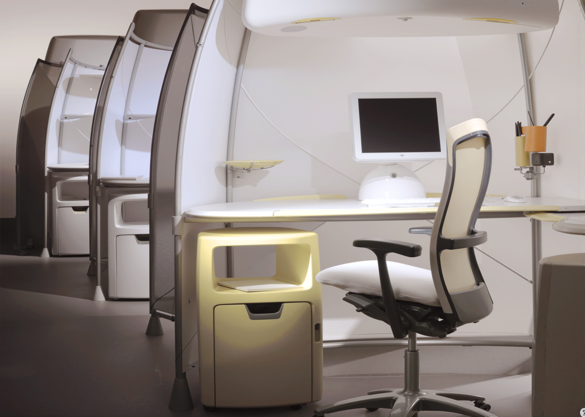 Interior view of a row of A3 workstations in grey showing the integrated desk with monitor, illuminated mobile storage cart, ergonomic chair, and overhead storage compartment