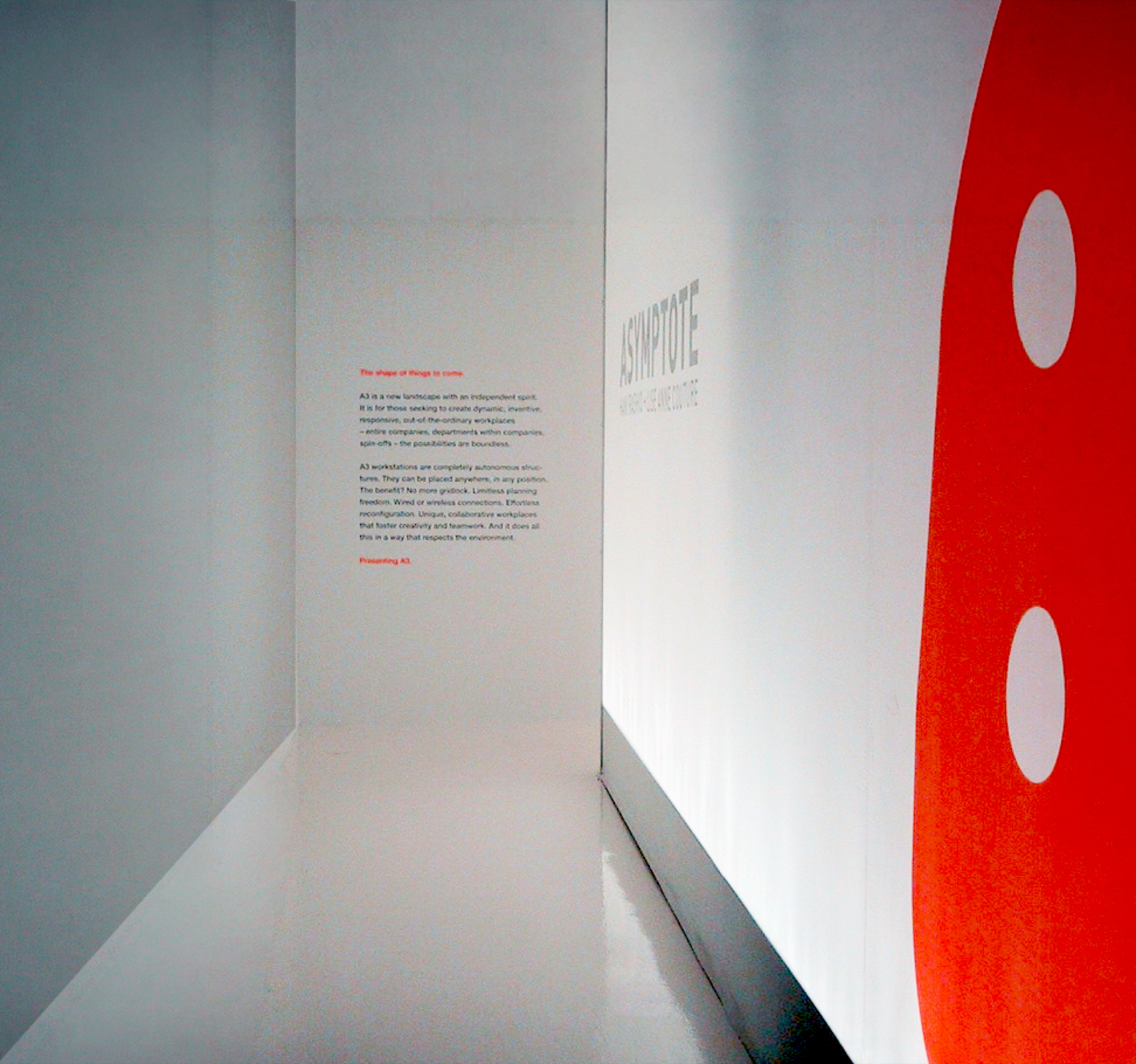 Narrow entry corridor with a curved red fabric wall and text panel introducing the A3 system concept, white surfaces framing the approach