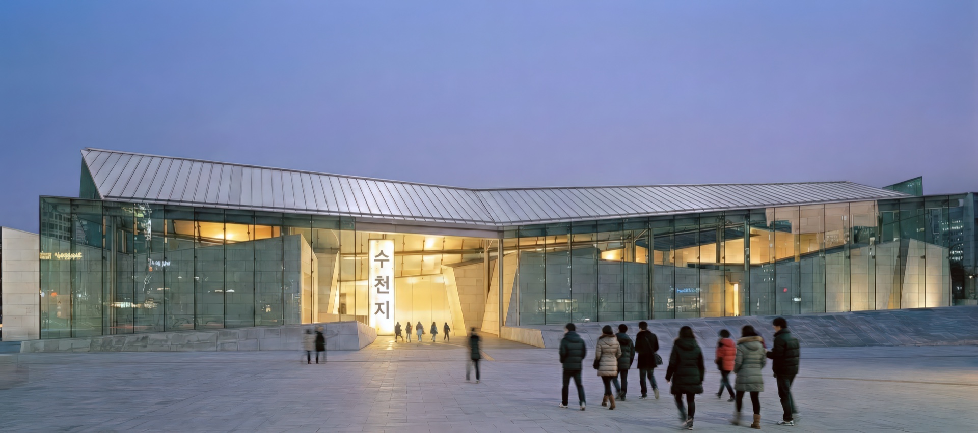 Panoramic view of the Korean History Museum entrance at twilight with visitors approaching the illuminated glass and stone pavilion