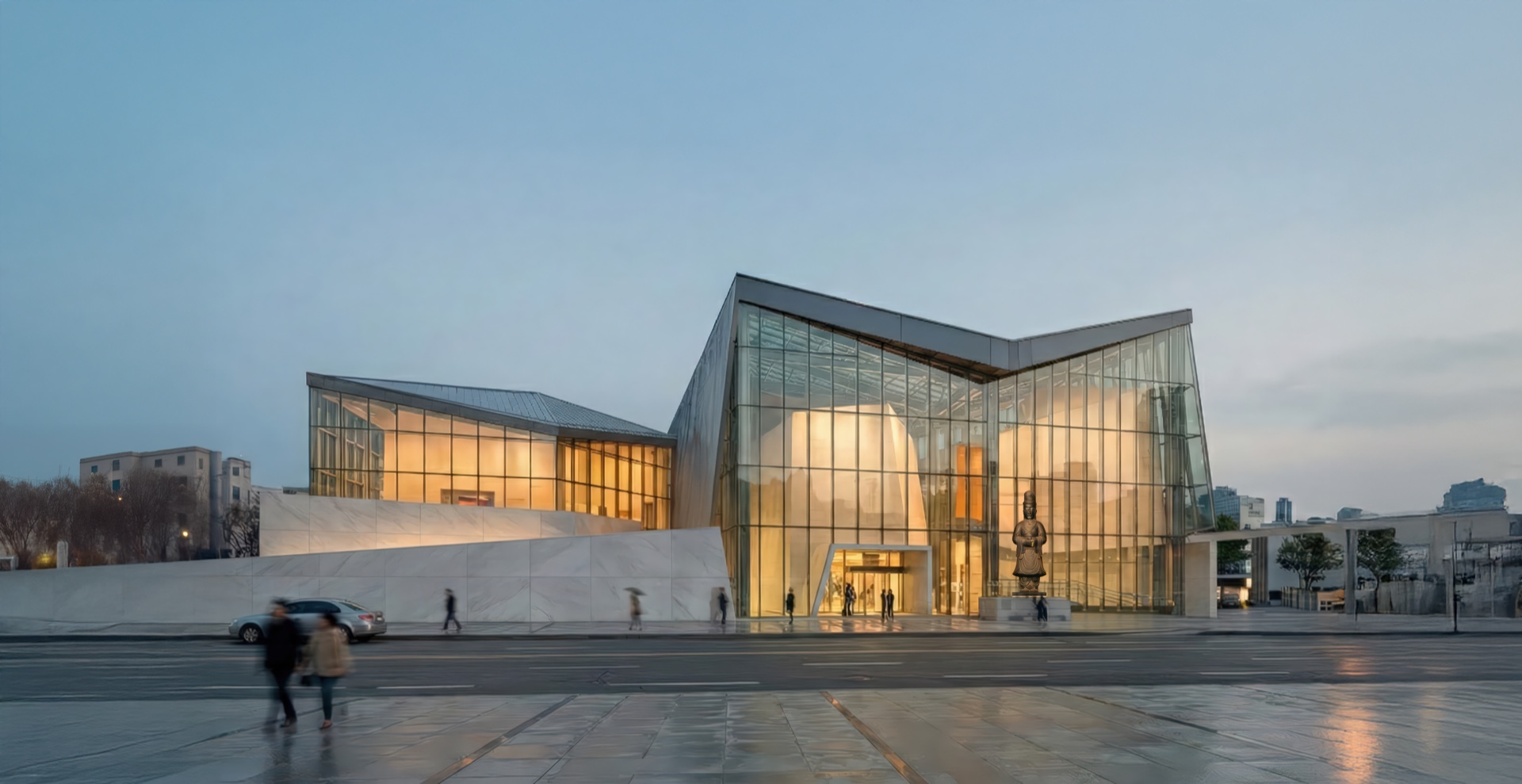 Exterior view of the Korean History Museum at dusk with its luminous glass facade and angular stone base glowing against the twilight sky