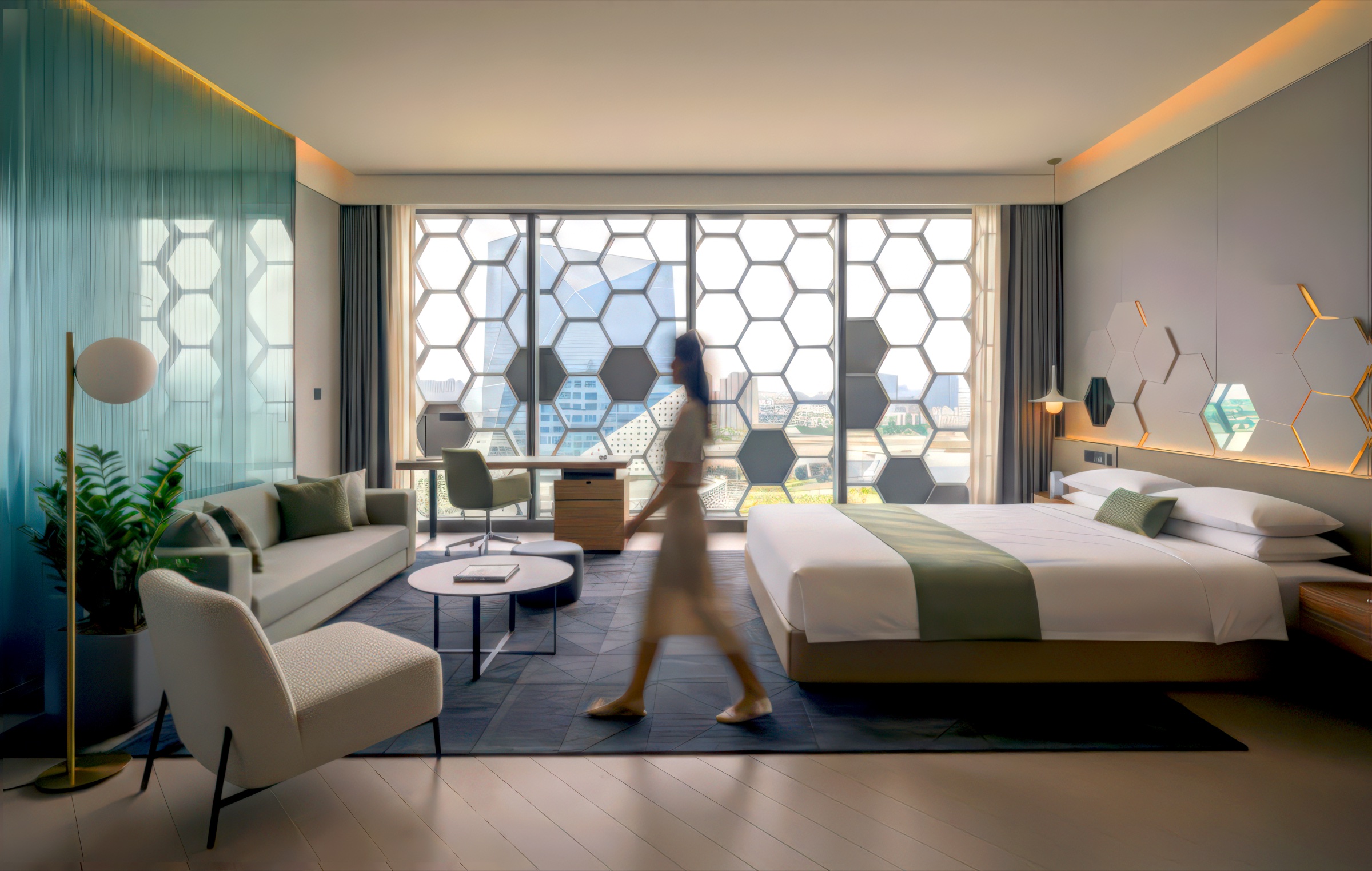 Deluxe guest room with hexagonal window wall filtering daylight, olive and white color palette, sofa seating area, work desk, and a guest walking through the space