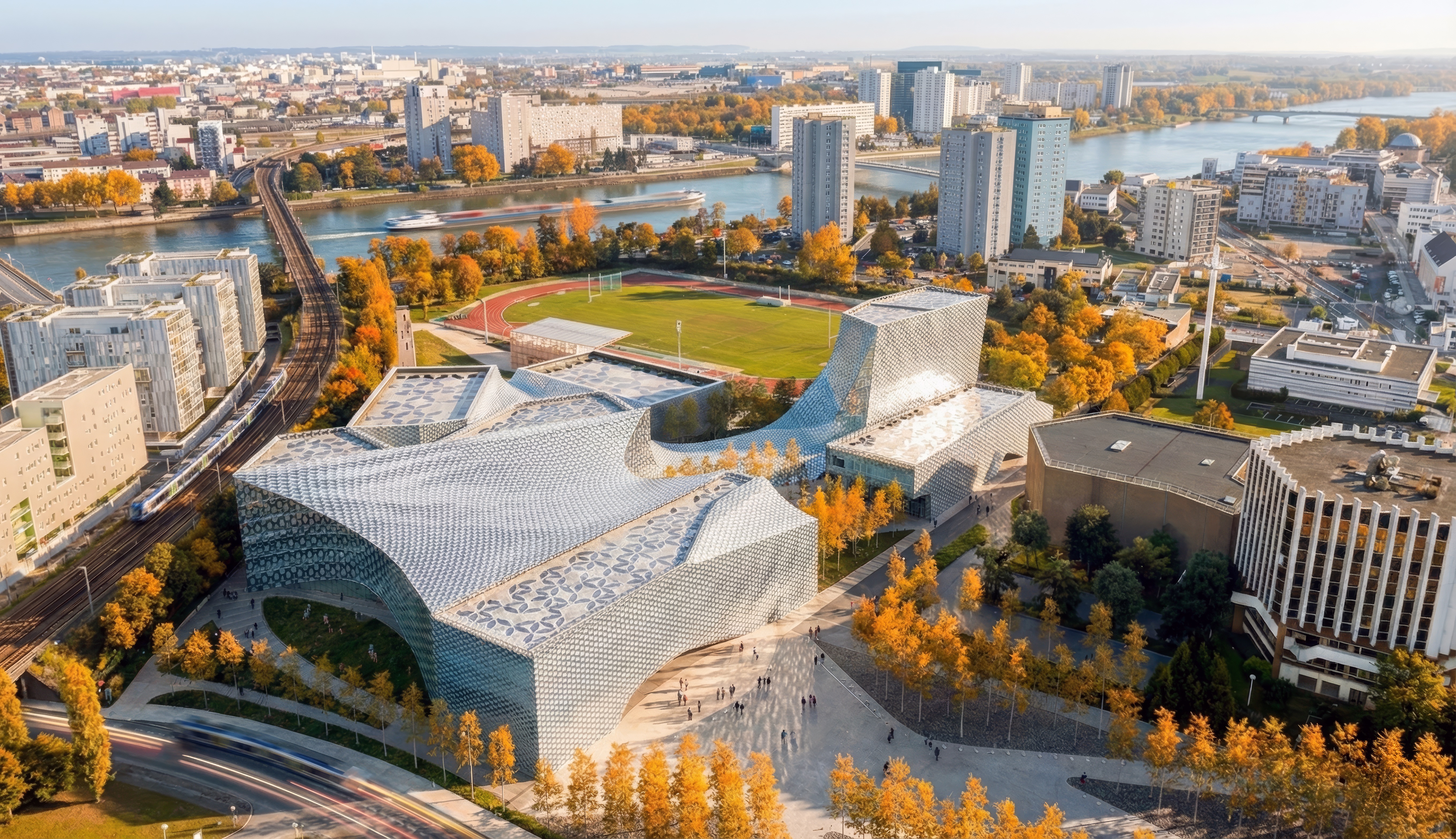 Aerial view of the Lycée Ile de Nantes campus set among autumn trees along the Loire River, showing the sculptural tessellated roof forms and adjacent sports fields