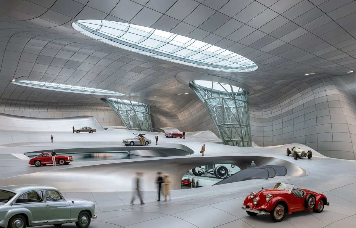 Wide interior view of the main exhibition hall with classic Mercedes vehicles arranged on a sculpted white floor topography, oval skylights overhead, and two diagrid-cage vertical lanterns drawing daylight into the space