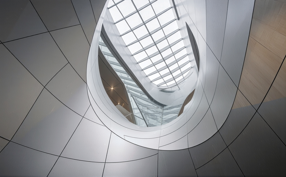 Low upward view of a large elliptical glazed roof oculus, the curved aluminium-clad ceiling surfaces sweeping up to frame the daylight aperture