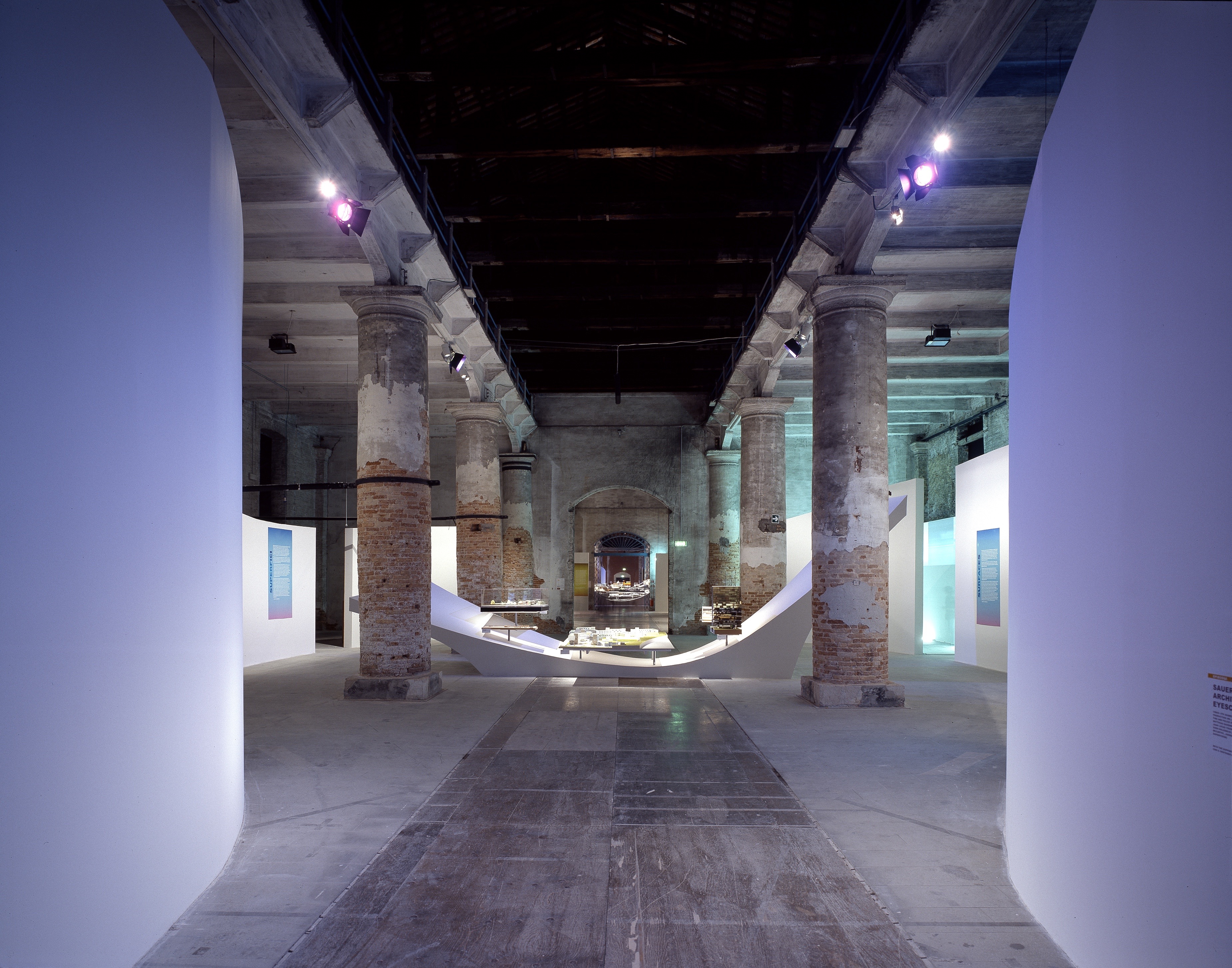 Deep perspective through the Corderie dell'Arsenale — blue-lit space with sweeping white display surfaces, architectural models, and historic stone columns receding into depth