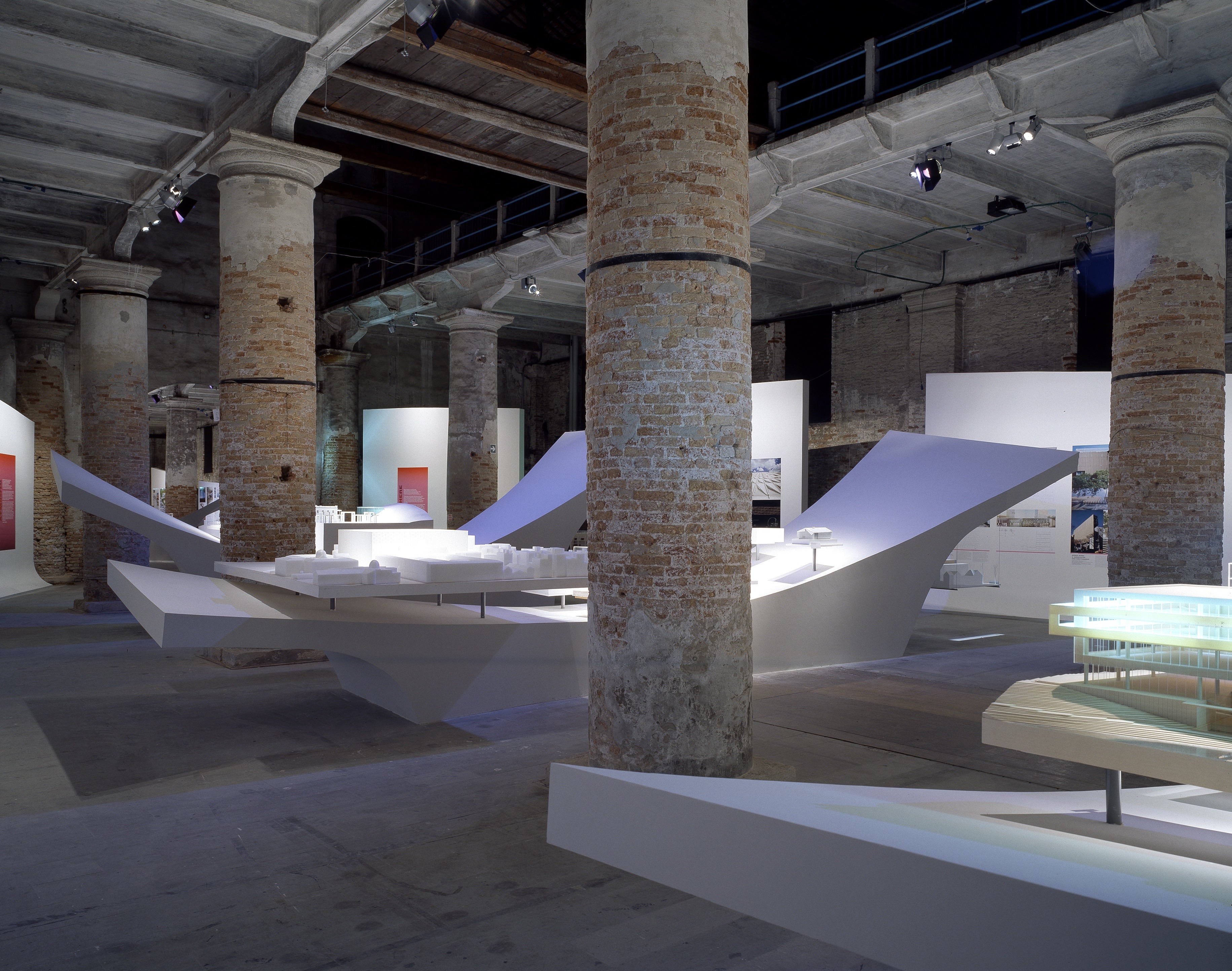 Curving white display platforms with architectural models set against exposed brick columns and blue-tinted lighting