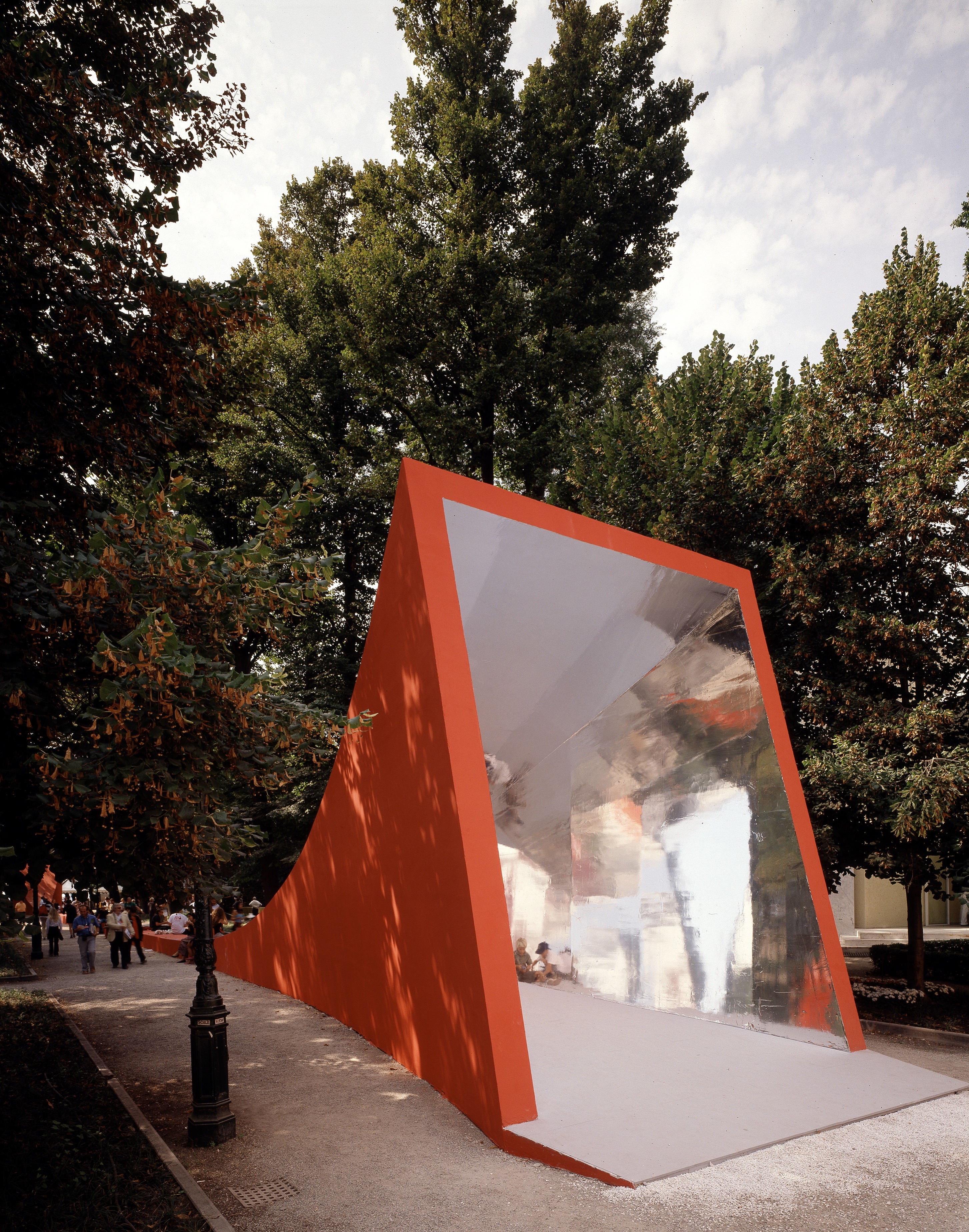 The Metamorph gateway pavilion in the Giardini — a bold red angular volume rising from the pavement among the trees, its open mirrored interior framing visitors and the Biennale grounds beyond