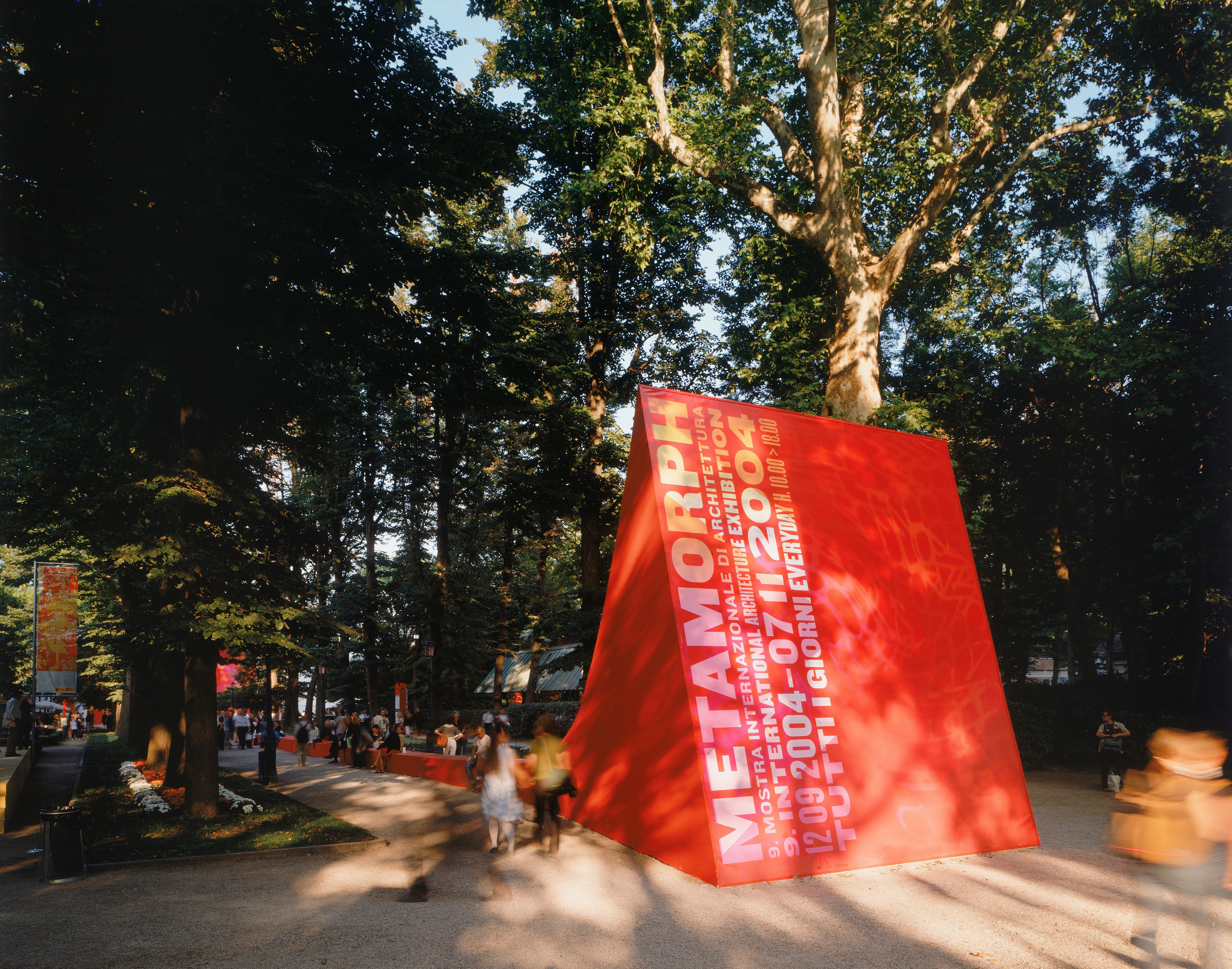 The entrance face of the Metamorph Giardini pavilion — bold red surfaces with gold typography displaying the Biennale graphic identity, visitors passing among the trees