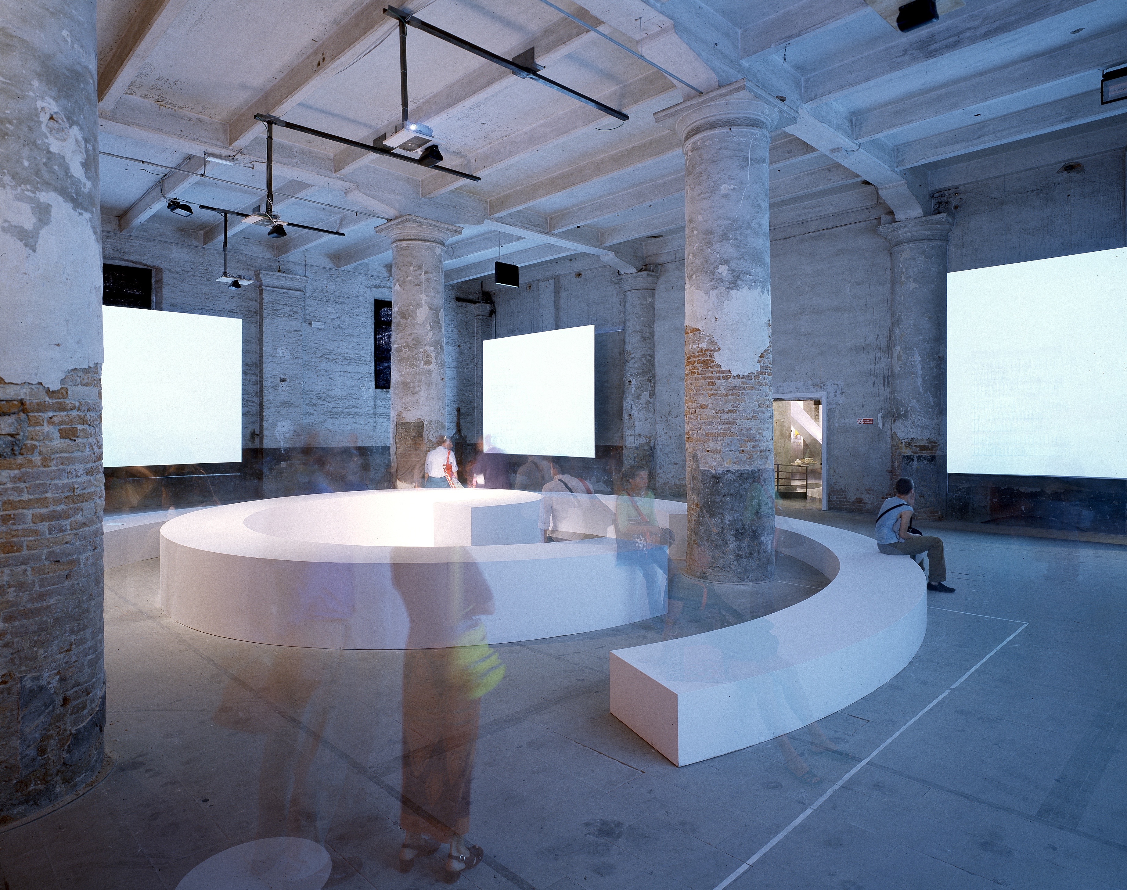 Circular white seating element with video projections in the Arsenale — visitors gathered among the columns