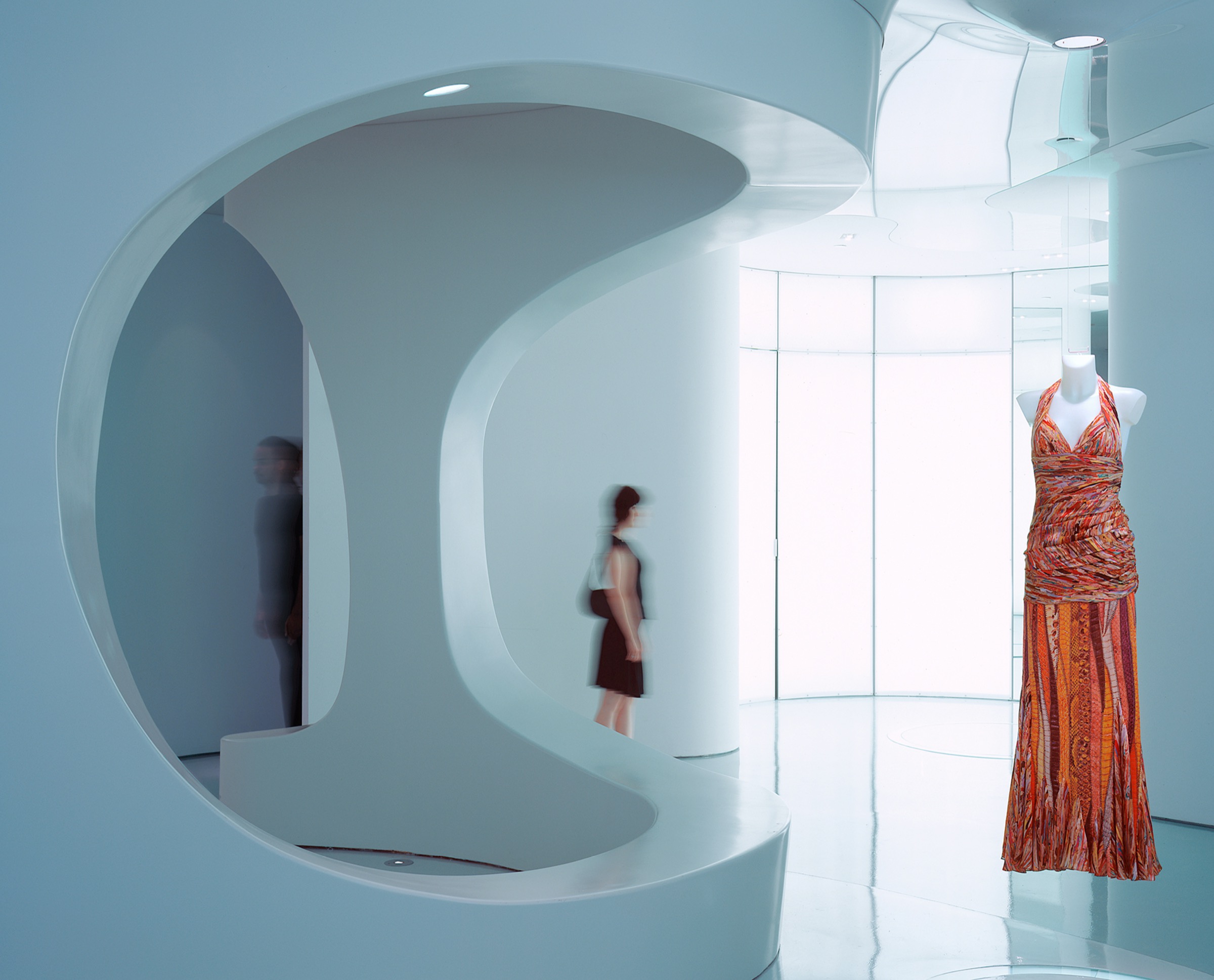 Interior view through a large curvilinear wall opening framing a motion-blurred visitor, an orange patterned dress on a mannequin, and luminous curved surfaces beyond